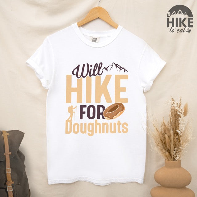 White Will Hike for Doughnuts statement t-shirt featuring chocolate doughnut graphic, mountain peaks, and hiker silhouette for doughnut lovers and outdoor enthusiasts on Comfort Colors 1717 hanging on wooden hanger