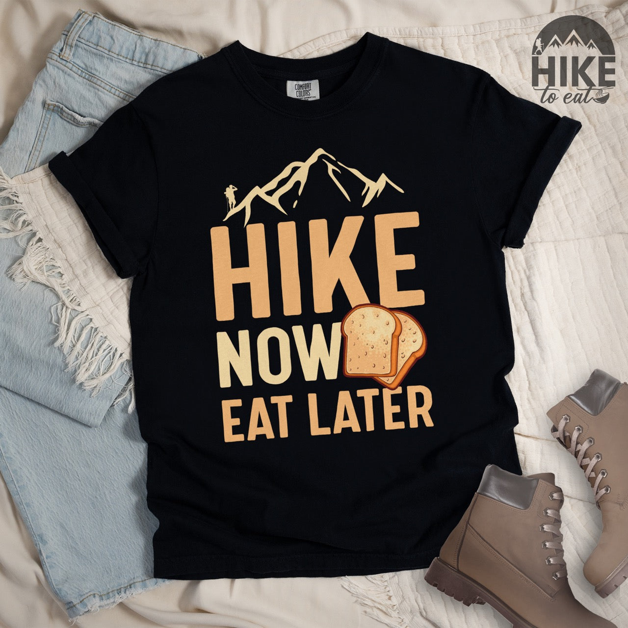 Black Food motivation vintage tee Hike Now Eat Later with mountain peak range character silhouette bold typography and illustrated toast slices on Comfort Colors shirt for delayed gratification adventure enthusiasts, food lovers and hikers - Laid flat with boots and pants.