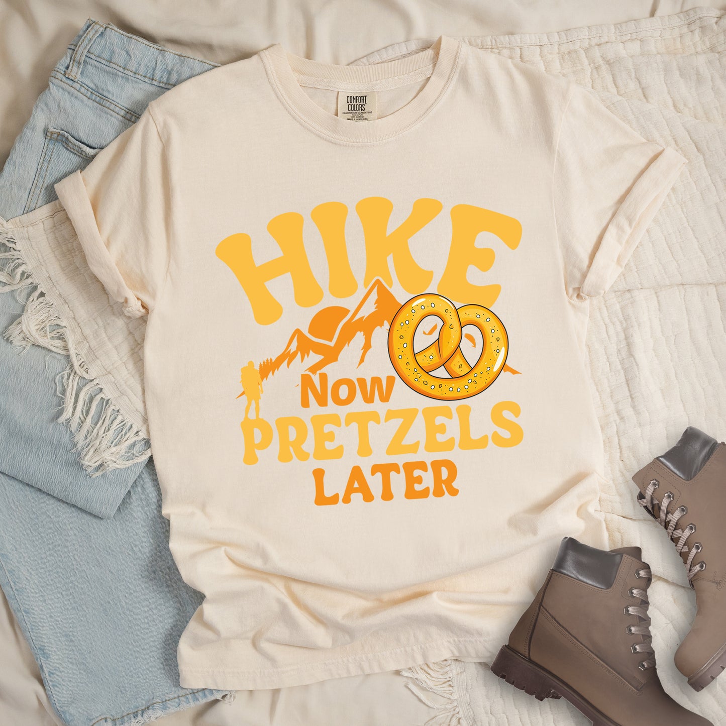 Ivory (Cream White) t-shirt with "HIKE NOW PRETZELS LATER" text design. "HIKE" appears at the top in yellow letters, "NOW" in orange, "PRETZELS" in yellow, and "LATER" in orange. The design includes orange mountain silhouettes with a small yellow hiker figure and an illustration of a golden pretzel with salt. Features a classic crew neck and casual rolled sleeves, styled alongside light wash denim and brown hiking boots.