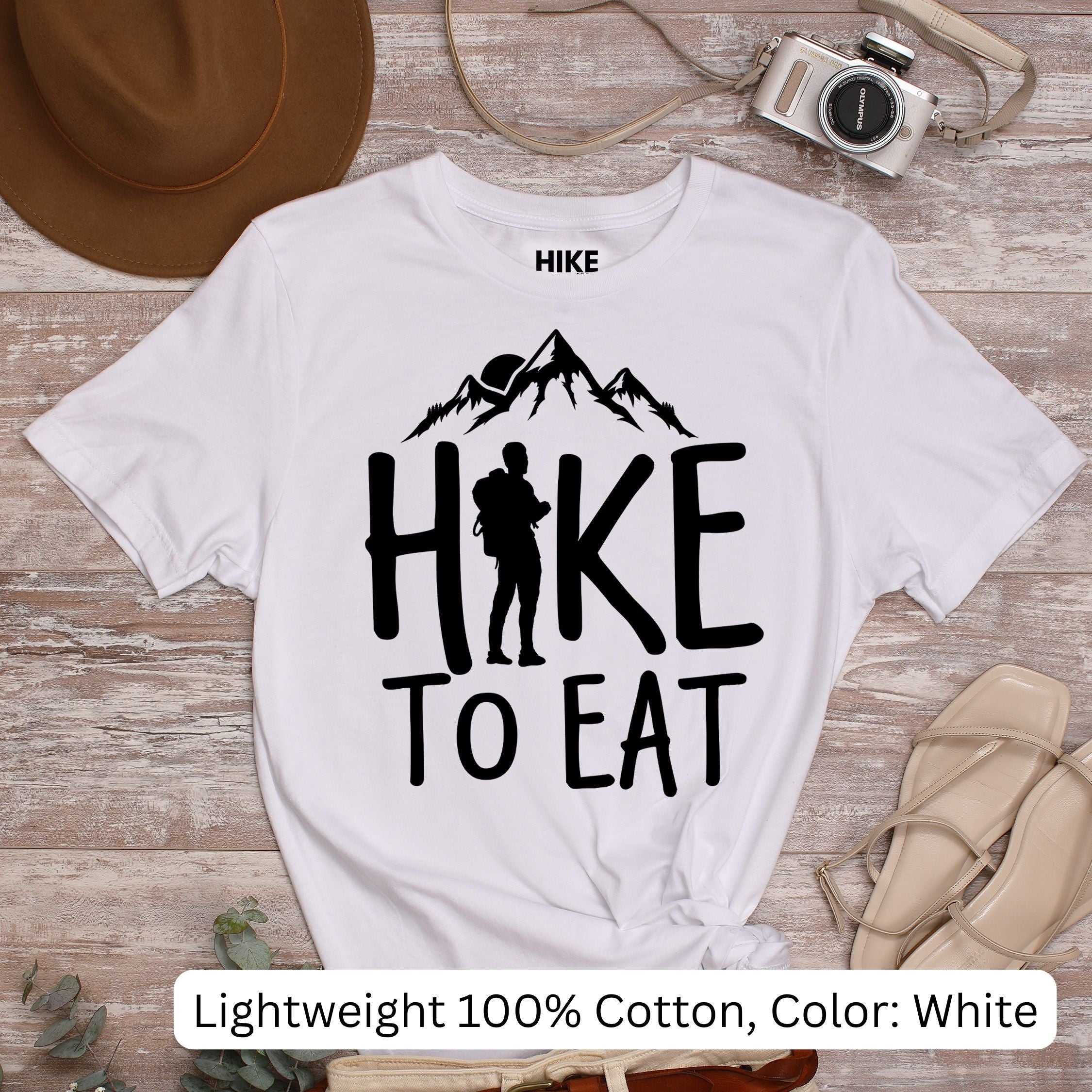 White t-shirt with 'Hike to Eat' design on a wooden surface with accessories.
