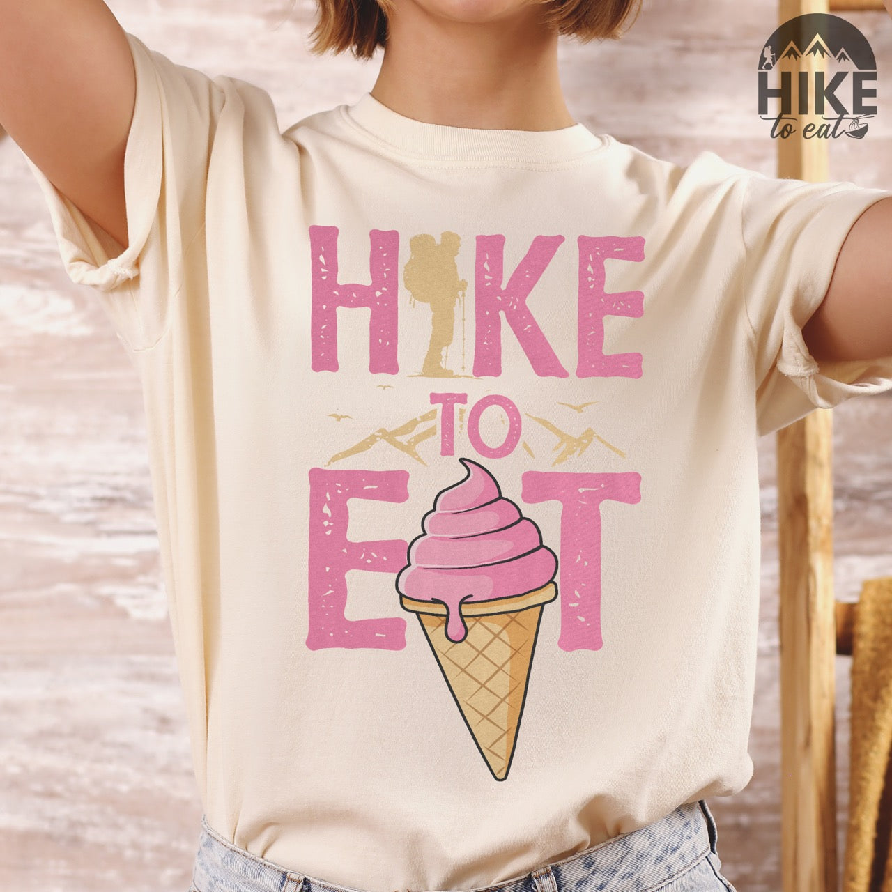 Ivory (beige) Hike to Eat brand logo t-shirt with distressed pink typography, hiker silhouette integrated into letters, mountain peaks, flying birds, and soft-serve ice cream cone illustration for dessert-loving outdoor enthusiasts