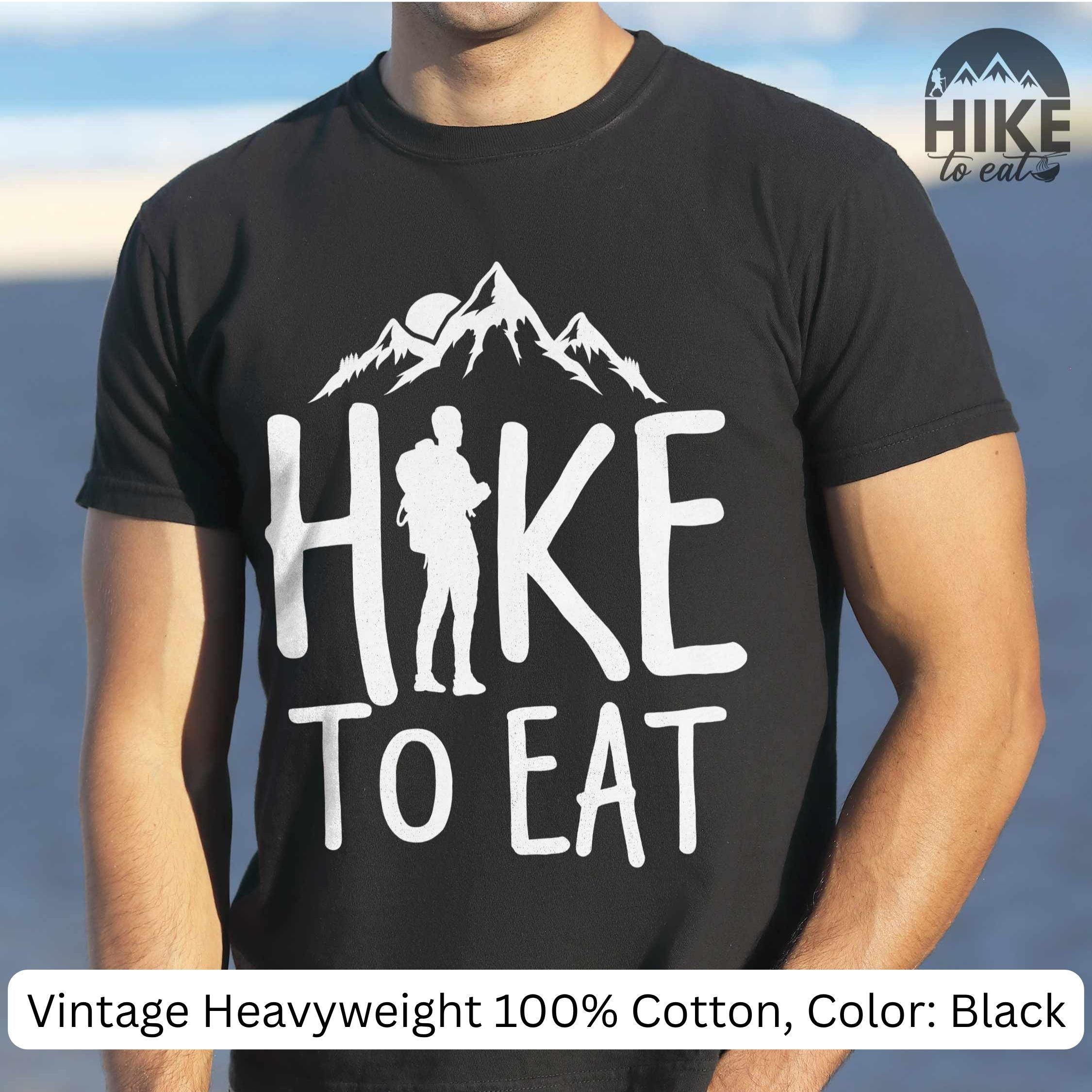 Black t-shirt with 'Hike to Eat' design worn by a person, with a mountainous background.