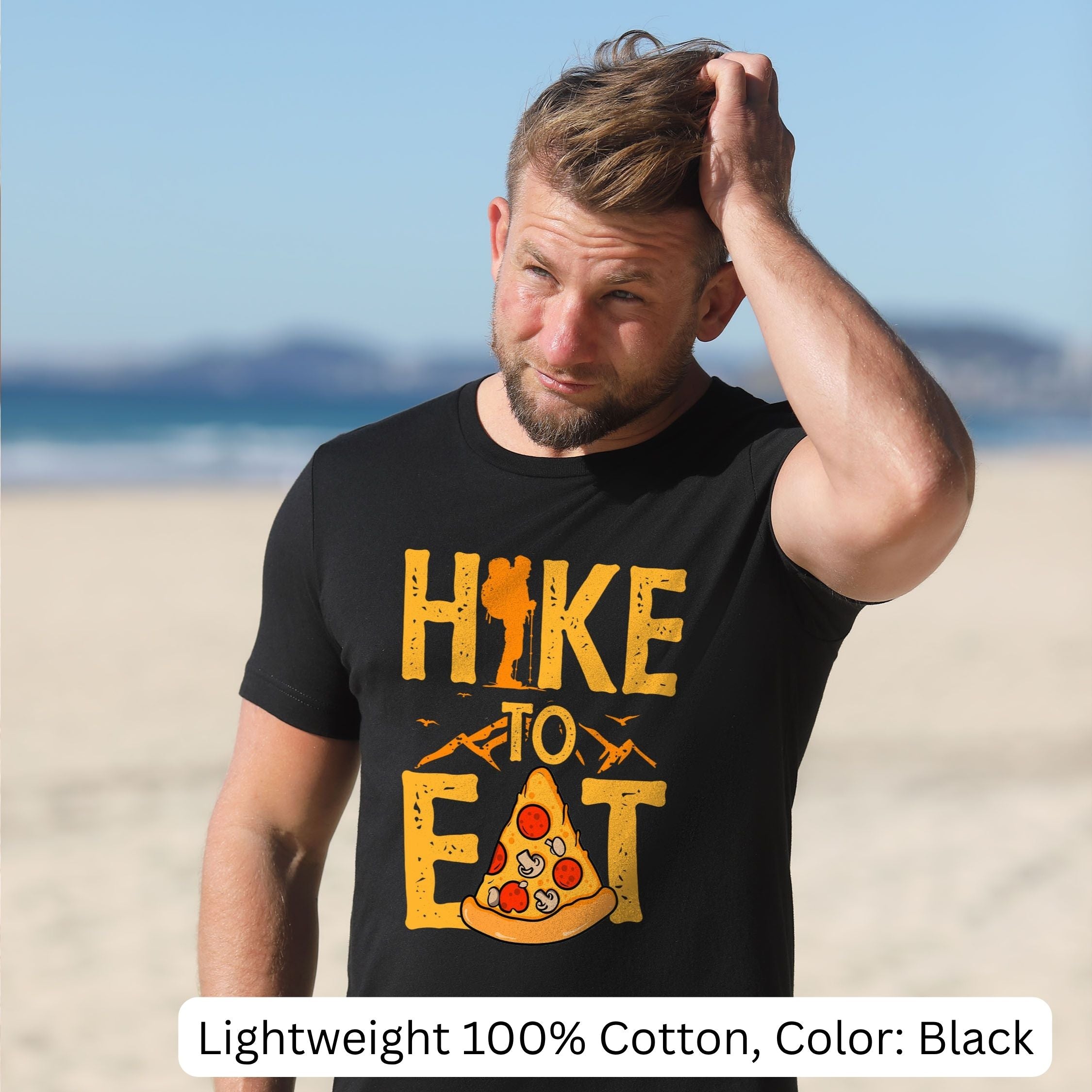 Man wearing a black t-shirt with 'Hike to Eat' design with the A as a pizza slice on a beach. Lightweight 100% cotton, Color: Black
