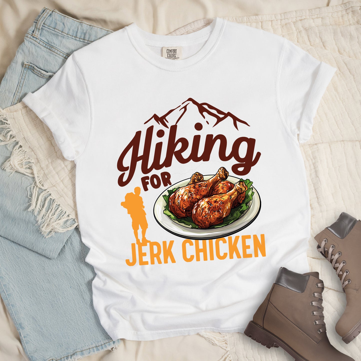 White t-shirt with brown and orange text reading "Hiking FOR JERK CHICKEN". "Hiking" appears in large brown script letters at the top with brown mountain outlines above it, "FOR" in brown text in the middle, and "JERK CHICKEN" in orange at the bottom. Features an illustration of jerk chicken drumsticks on a white plate with green garnish in the center, and a small orange hiker silhouette to the left of the chicken illustration and above the words "JERK CHICKEN".