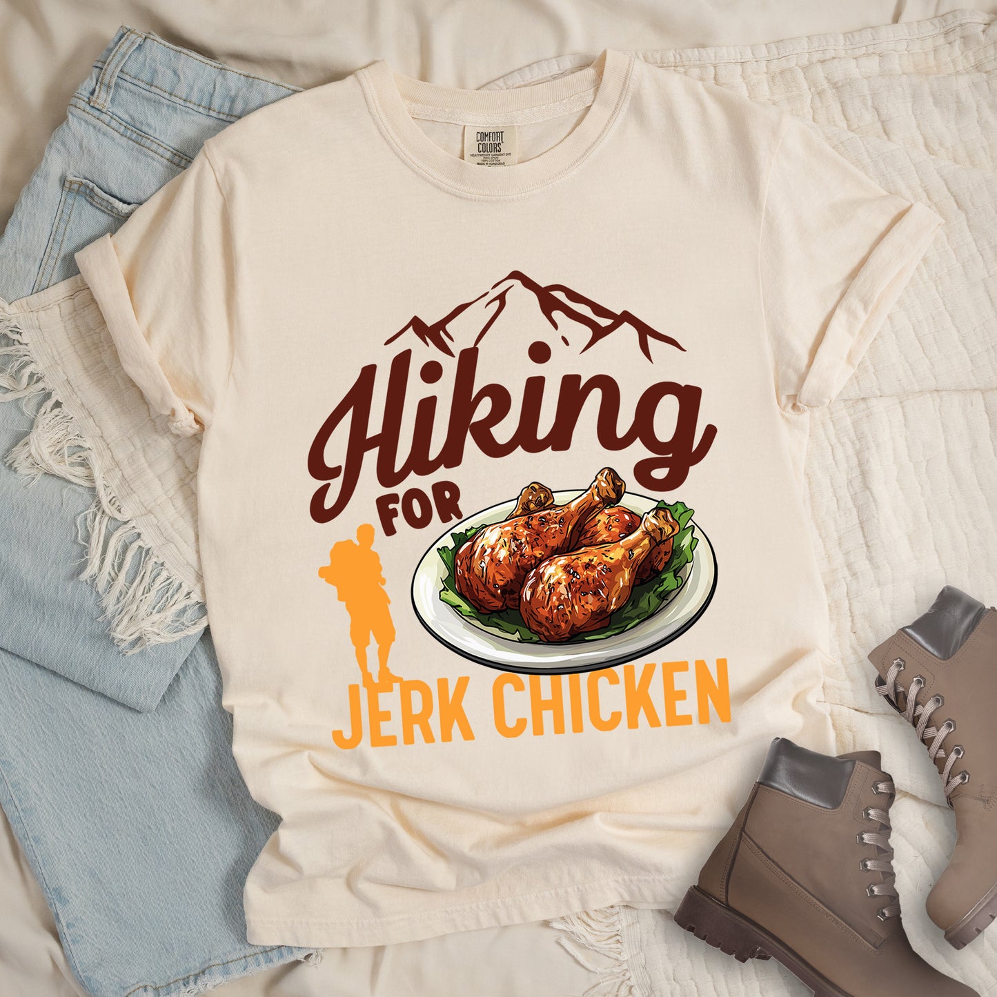 Ivory (Cream White) t-shirt with brown and orange text reading "Hiking FOR JERK CHICKEN". "Hiking" appears in large brown script letters at the top with brown mountain outlines above it, "FOR" in brown text in the middle, and "JERK CHICKEN" in orange at the bottom. Features an illustration of jerk chicken drumsticks on a white plate with green garnish in the center, and a small orange hiker silhouette to the left of the chicken illustration and above the words "JERK CHICKEN".