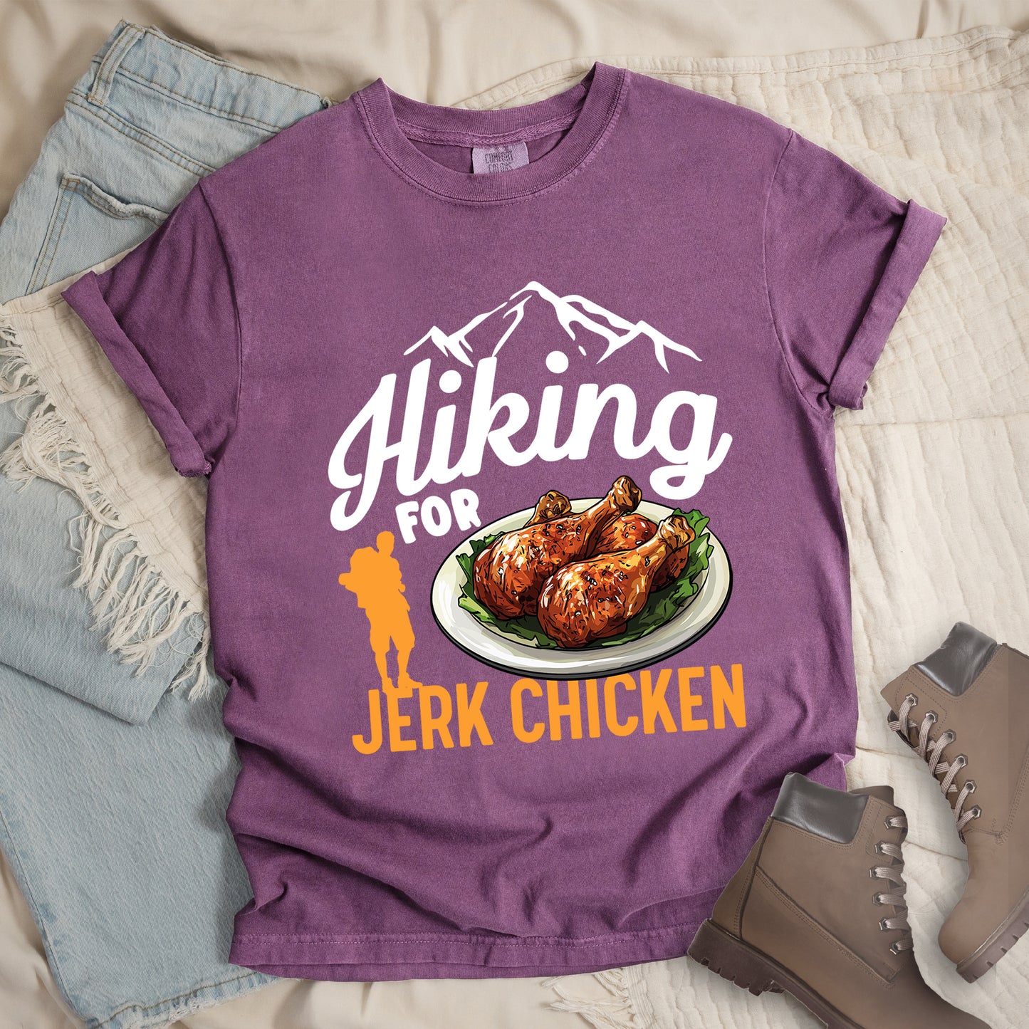 Berry (Dark Violet) t-shirt with white and orange text reading "Hiking FOR JERK CHICKEN". "Hiking" appears in large white script letters at the top with white mountain outlines above it, "FOR" in white text in the middle, and "JERK CHICKEN" in orange at the bottom. Features an illustration of jerk chicken drumsticks on a white plate with green garnish in the center, and a small orange hiker silhouette to the left of the chicken illustration and above the words "JERK CHICKEN".