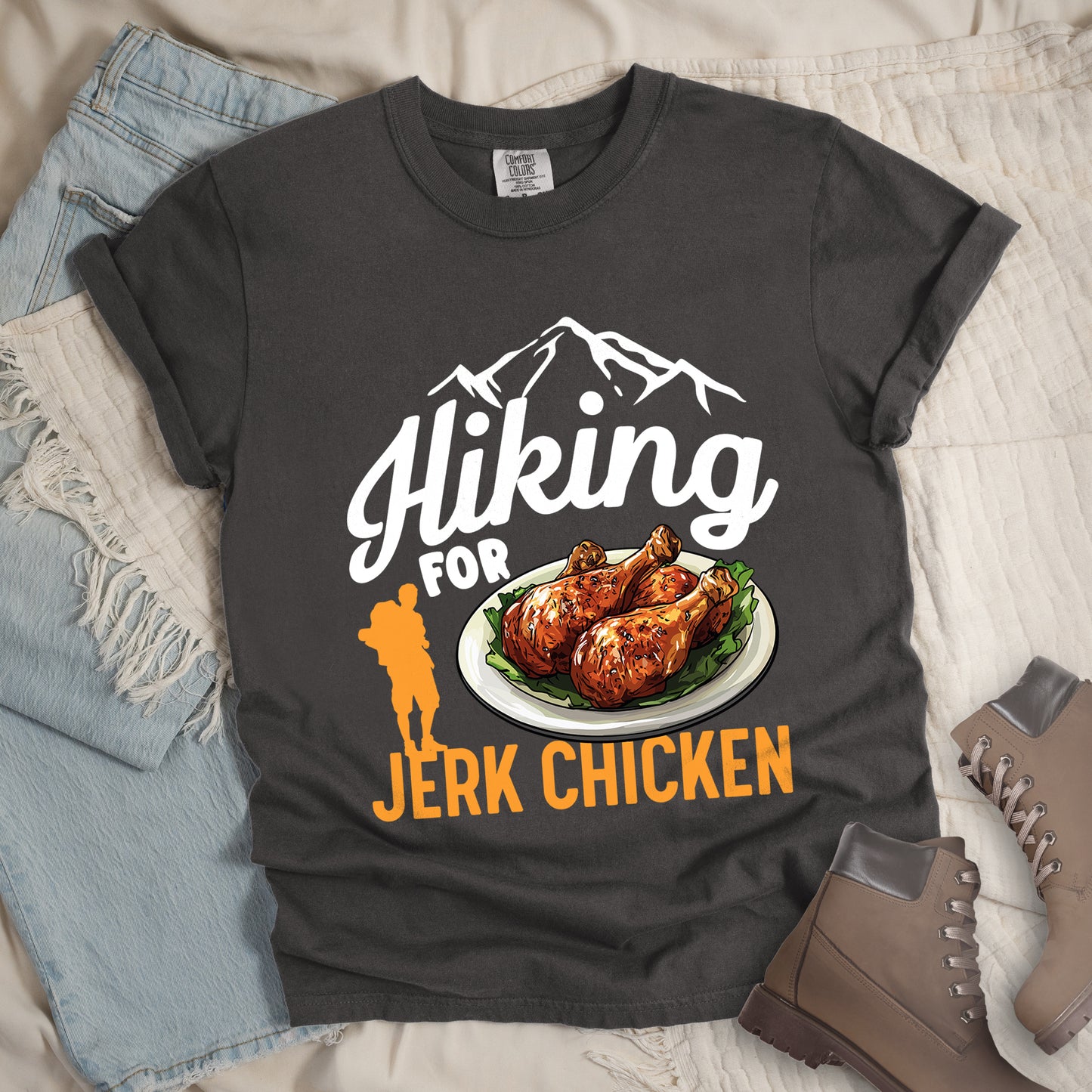 Pepper (Dark Gray) t-shirt with white and orange text reading "Hiking FOR JERK CHICKEN". "Hiking" appears in large white script letters at the top with white mountain outlines above it, "FOR" in white text in the middle, and "JERK CHICKEN" in orange at the bottom. Features an illustration of jerk chicken drumsticks on a white plate with green garnish in the center, and a small orange hiker silhouette to the left of the chicken illustration and above the words "JERK CHICKEN".