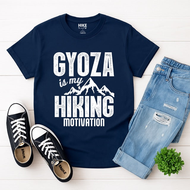 Navy blue Gyoza is my Hiking Motivation statement t-shirt with bold distressed typography, mountain peaks, and hiker silhouette for gyoza lovers and outdoor enthusiasts on Softstyle 100% cotton with pants, shoes and plant next to it, laying flat.