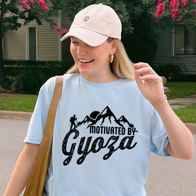 woman wearing light blue Dark Chocolate (brown) Motivated by Gyoza statement tee with distressed typography, mountain peaks, and hiker silhouette for gyoza lovers and japanese food lovers.