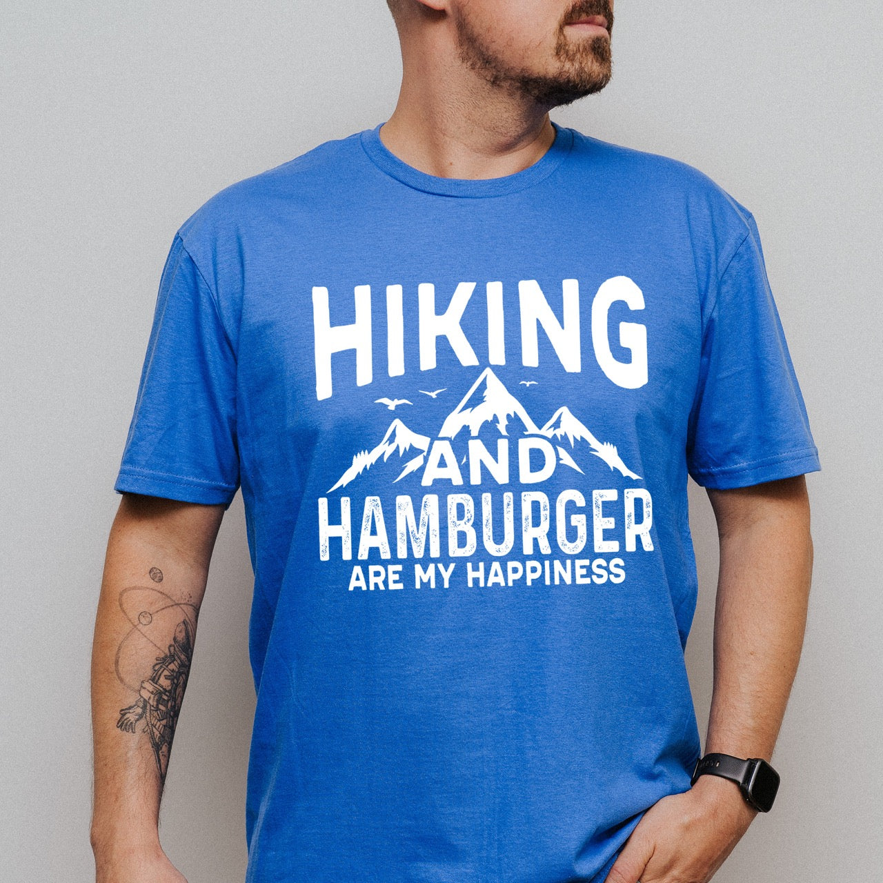 Man wearing a royal (blue) Hiking and Hamburger are my Happiness statement tee with distressed bold typography, mountain peaks, flying birds, and pine trees for burger-loving outdoor enthusiasts and hikers