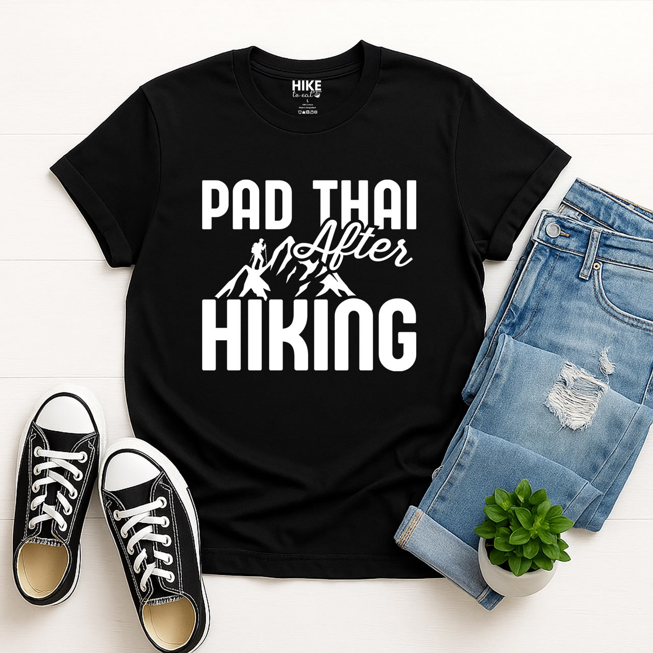 black Pad Thai after Hiking statement tee with bold typography, elegant script lettering, mountain peaks, and hiker silhouette for Thai food-loving outdoor enthusiasts, foodies and hikers, pictured with jeans and shoes