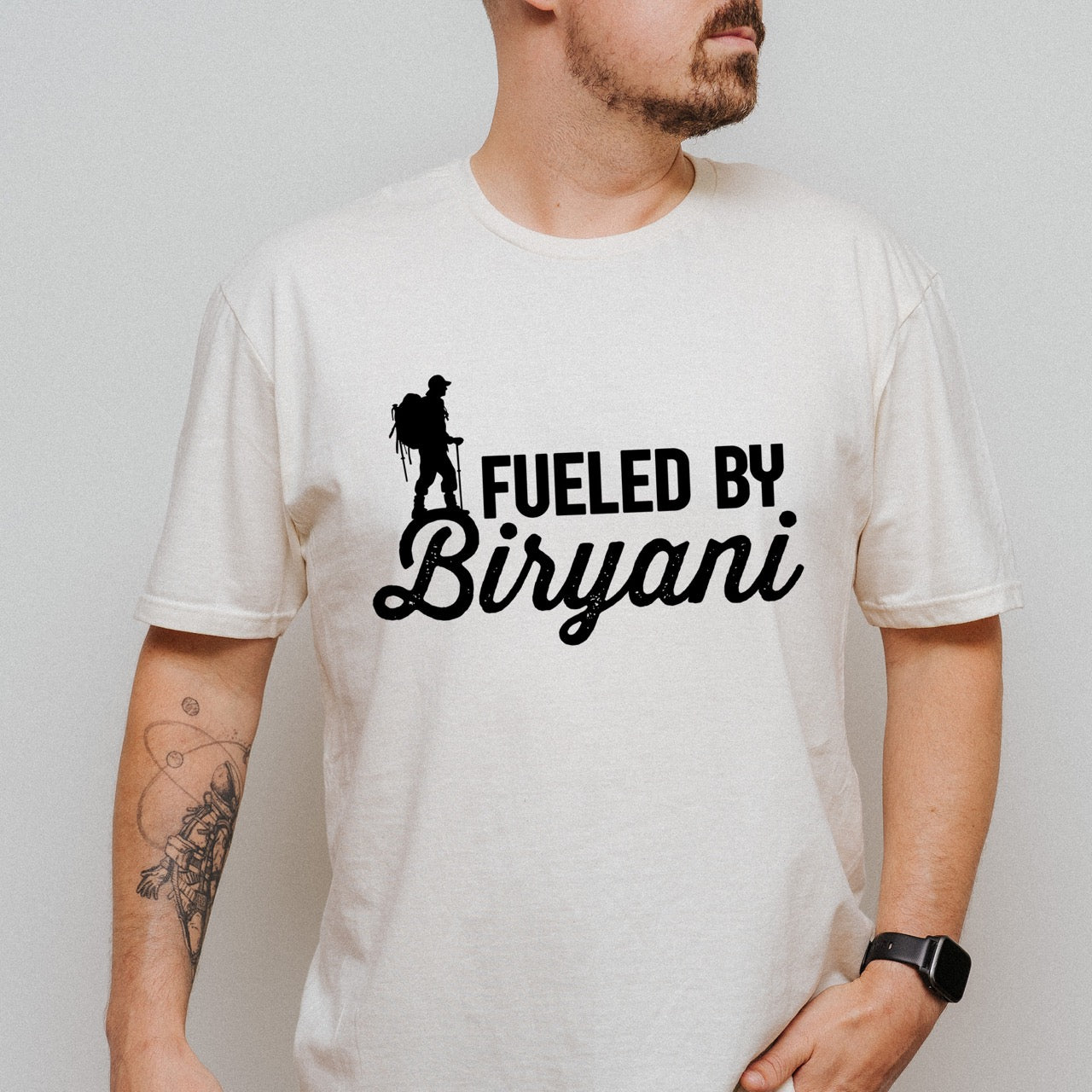 woman wearing Natural (beige) Fueled by Biryani statement tee with bold typography, elegant script lettering, and hiker silhouette for Indian food-loving outdoor enthusiasts, foodies and hikers