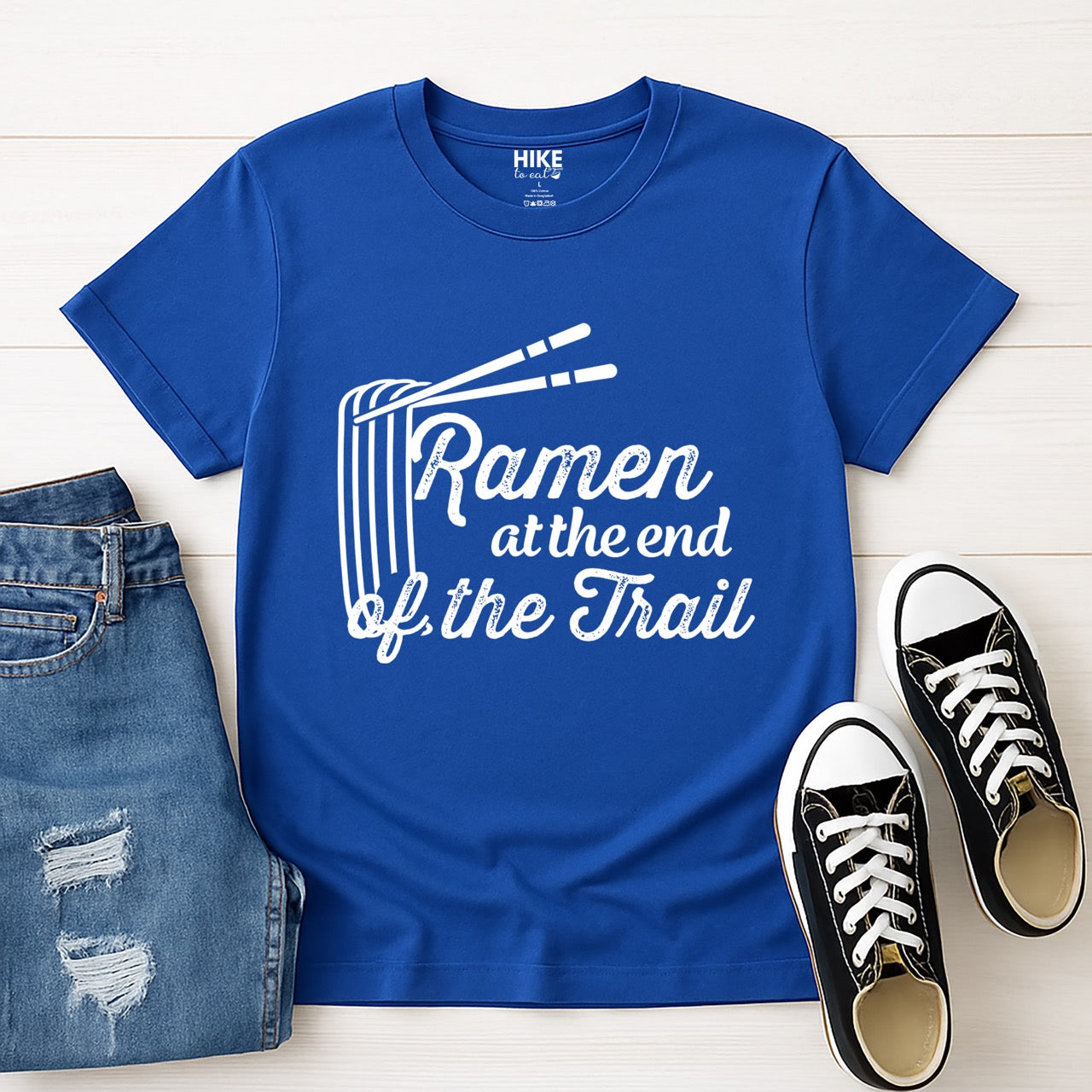 Royal blue Ramen at the End of the Trail statement tee with elegant script typography, ramen bowl illustration, and chopsticks for Japanese noodle-loving outdoor enthusiasts, foodies and hikers laid flat with shoes and jeans