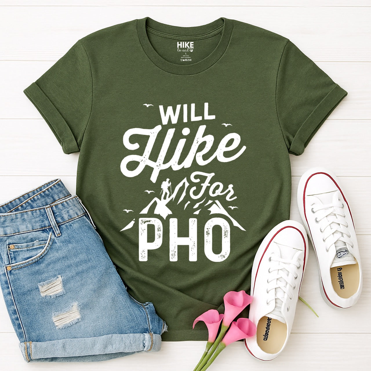 military green Will Hike for Pho statement tee with bold and script typography, mountain peaks, hiker silhouette, and flying birds for Vietnamese noodle-loving outdoor enthusiasts, hikers and foodies laid flat with jeans and shoes