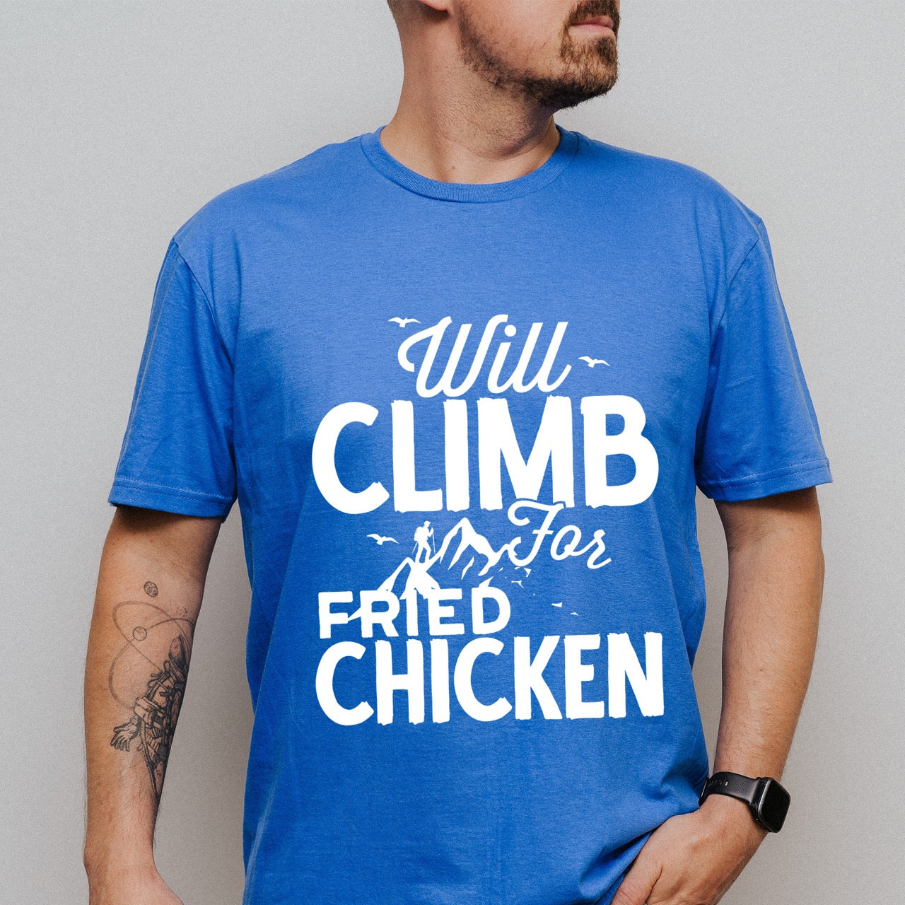 Man wearing Royal Blue Will climb for Fried Chicken statement tee with bold and script typography, mountain peaks, climber silhouette, and flying birds for chicken-loving outdoor athletes, climbers, hikers and foodies