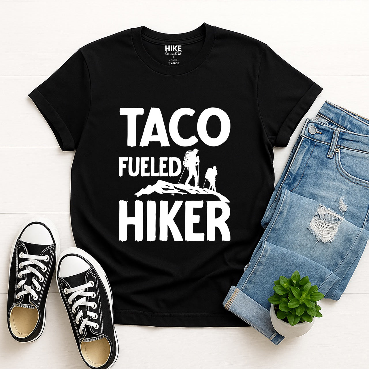 Black Taco Fueled Hiker statement tee with bold distressed typography and two hiker silhouettes on mountain ridge for Mexican food-loving outdoor enthusiasts, hikers and foodies laid flat with jeans and shoes