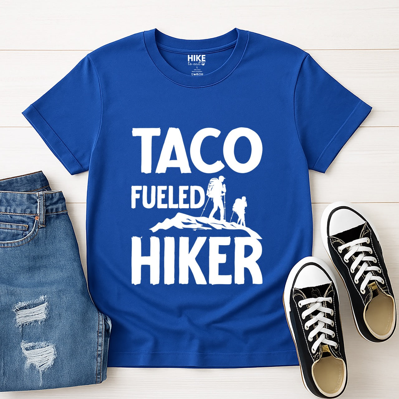 Royal blue Taco Fueled Hiker statement tee with bold distressed typography and two hiker silhouettes on mountain ridge for Mexican food-loving outdoor enthusiasts, hikers and foodies laid flat with shoes and jeans