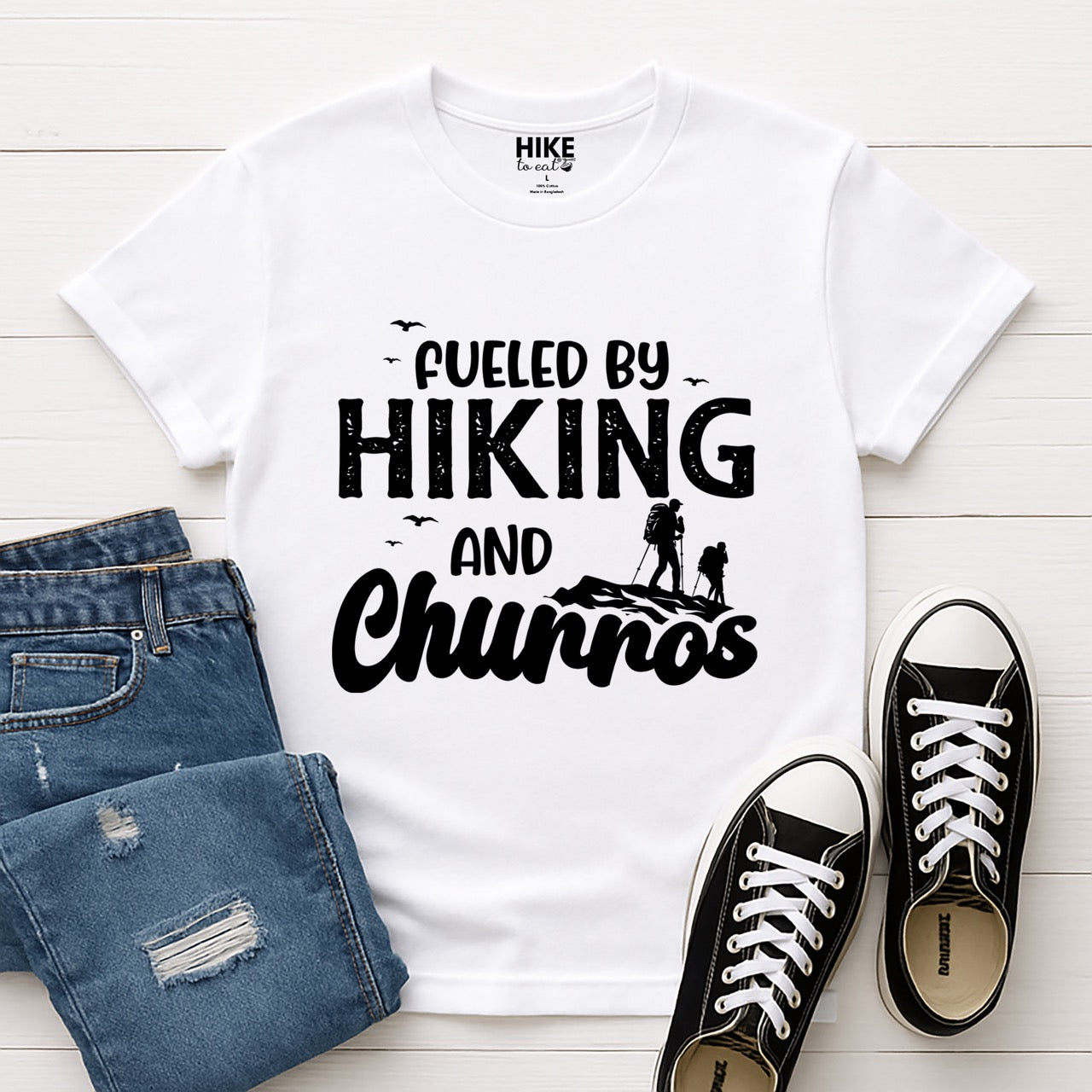 White Fueled by Hiking and Churros statement tee with bold distressed typography, flying birds, and two hiker silhouettes on mountain ridge for Spanish dessert-loving outdoor enthusiasts, hikers and foodies laid flat with shoes and pants