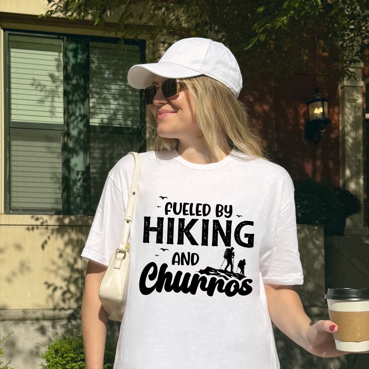 Woman wearing White Fueled by Hiking and Churros statement tee with bold distressed typography, flying birds, and two hiker silhouettes on mountain ridge for Spanish dessert-loving outdoor enthusiasts, hikers and foodies