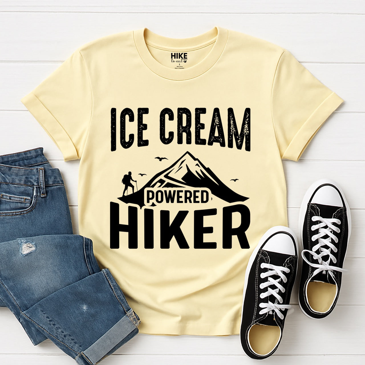 Cornsilk color (yellow) Ice Cream Powered Hiker statement tee with bold distressed typography, mountain peaks, hiker silhouette, and flying birds for dessert-loving outdoor enthusiasts, hikers and foodies laid flat with jeans and shoes