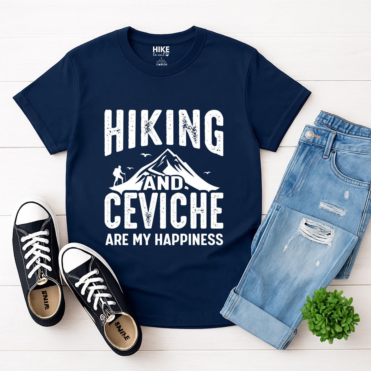Navy Hiking and Ceviche are my Happiness statement tee with distressed bold typography, mountain peaks, hiker silhouette, and flying birds for seafood-loving outdoor enthusiasts, foodies and hikers laid flat with pants and shoes