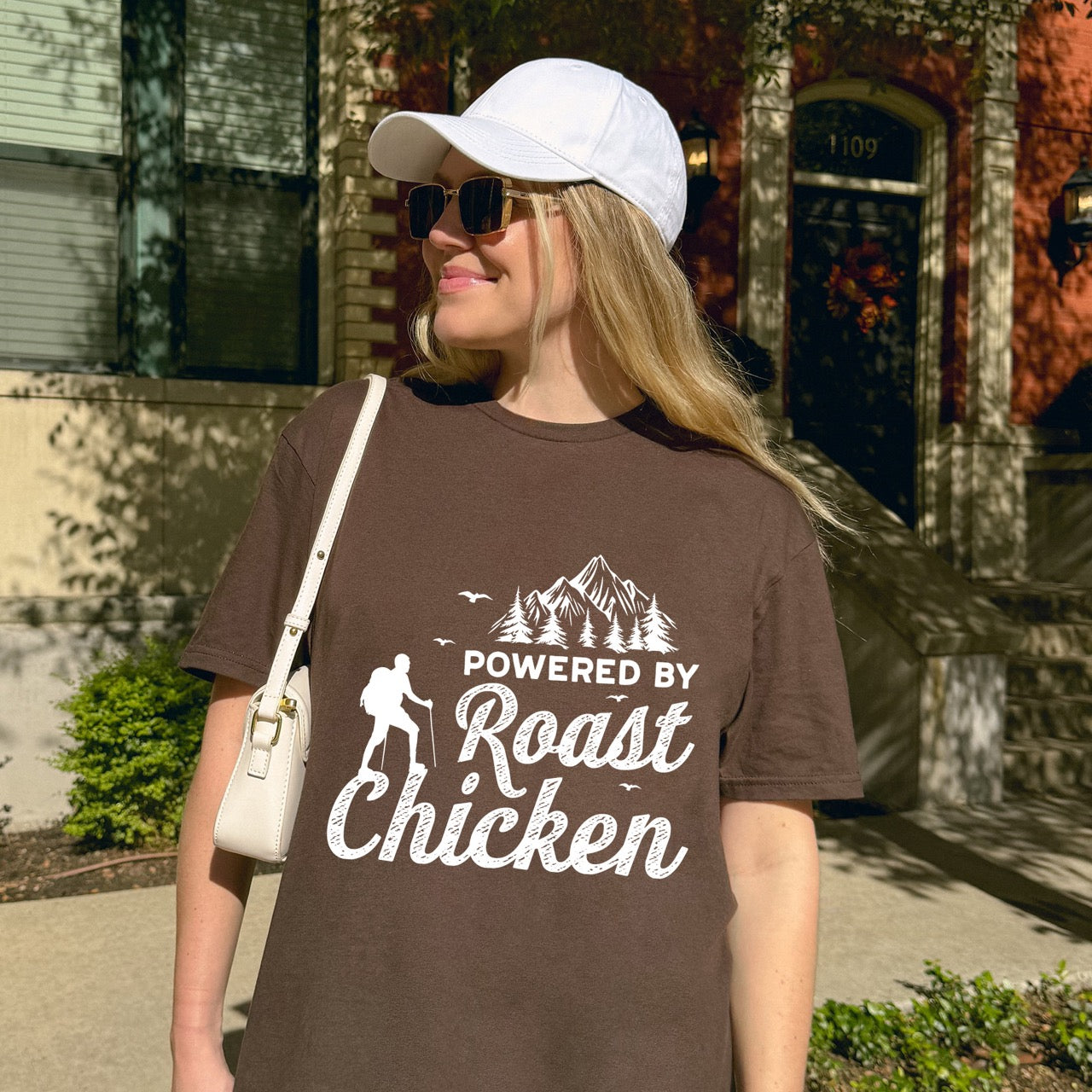 woman wearing dark chocolate Powered by Roast Chicken statement tee with bold typography, distressed script lettering, mountain peaks, pine trees, hiker silhouette, and flying birds for chicken-loving outdoor enthusiasts, hikers and foodies