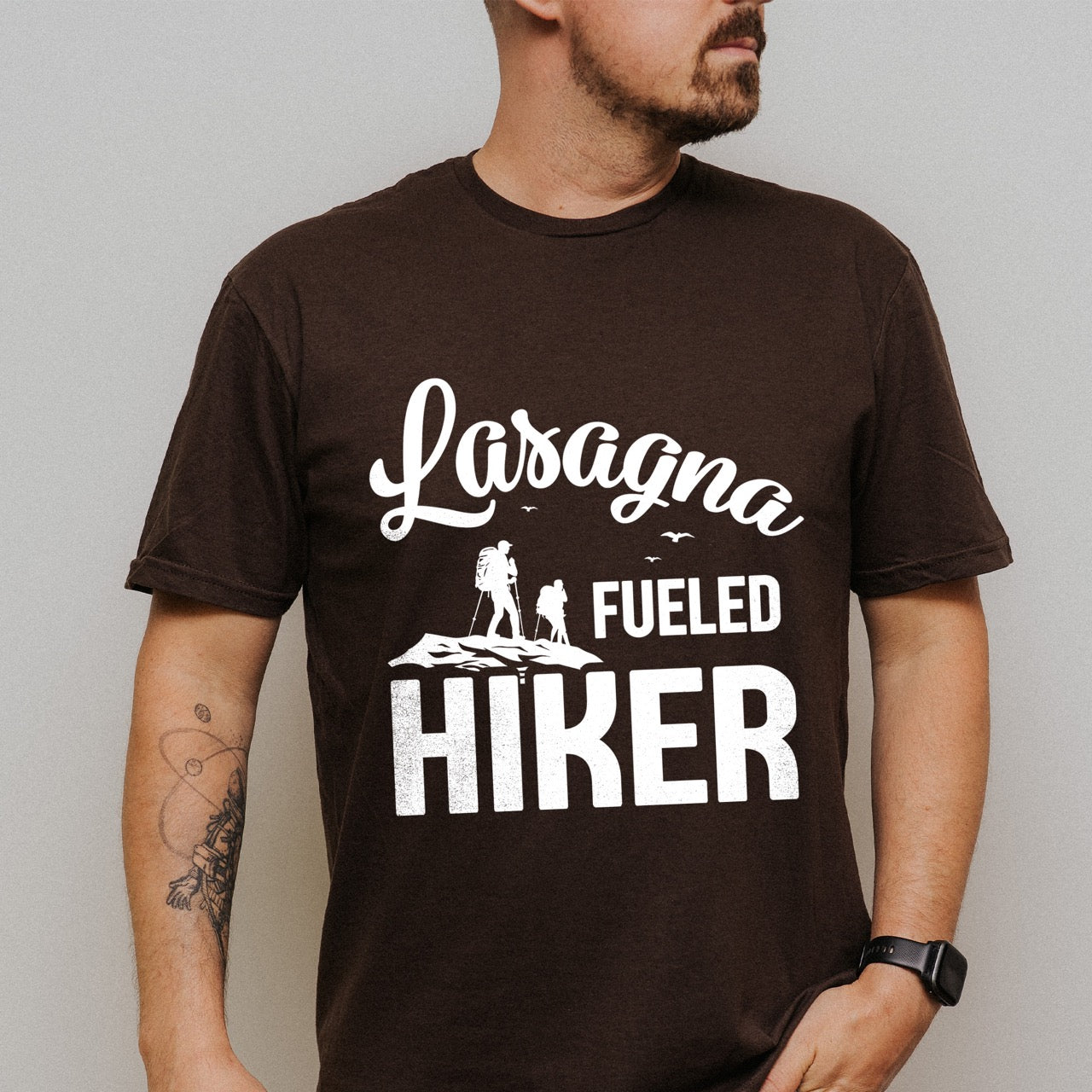 Man wearing Dark Chocolate (brown) Lasagna Fueled Hiker statement tee with flowing script typography, bold lettering, two hiker silhouettes on mountain ridge, and flying birds for Italian food-loving outdoor enthusiasts, hikers and foodies