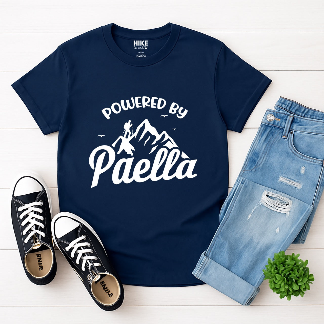 Navy Powered by Paella statement tee with bold curved typography, flowing script lettering, mountain peaks, hiker silhouette, and flying birds for Spanish food-loving outdoor enthusiasts, hikers and foodies laid flat with shoes and pants