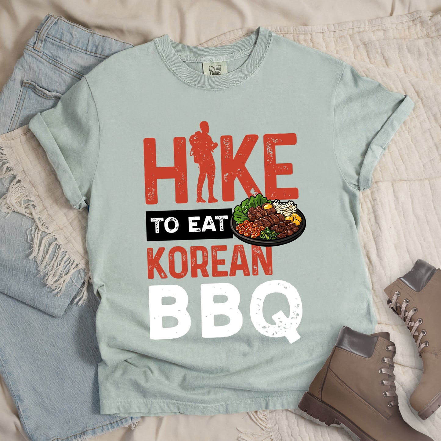 Bay (Light Gray) t-shirt with red and white distressed text reading "HIKE TO EAT KOREAN BBQ". "HIKE" appears in large red letters at the top with a red hiker silhouette integrated into the letter "I", "TO EAT" in white text on a black rectangular background in the middle, "KOREAN" in red, and "BBQ" in white at the bottom. Features an illustration of Korean barbecue with meat and vegetables on a black plate extending from the black background.