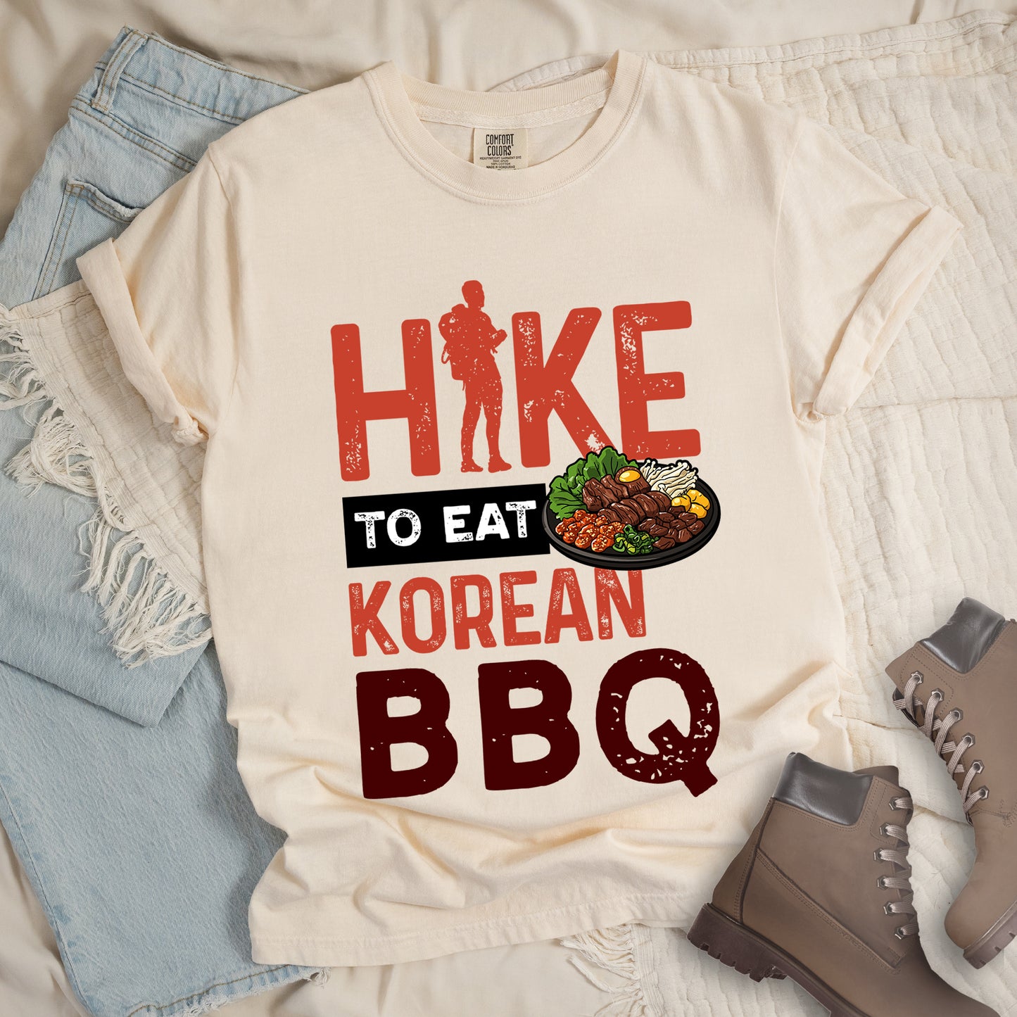 Ivory (Cream White) t-shirt with red and dark red distressed text reading "HIKE TO EAT KOREAN BBQ". "HIKE" appears in large red letters at the top with a red hiker silhouette integrated into the letter "I", "TO EAT" in white text on a black rectangular background in the middle, "KOREAN" in red, and "BBQ" in dark red at the bottom. Features an illustration of Korean barbecue with meat and vegetables on a black plate extending from the black background.
