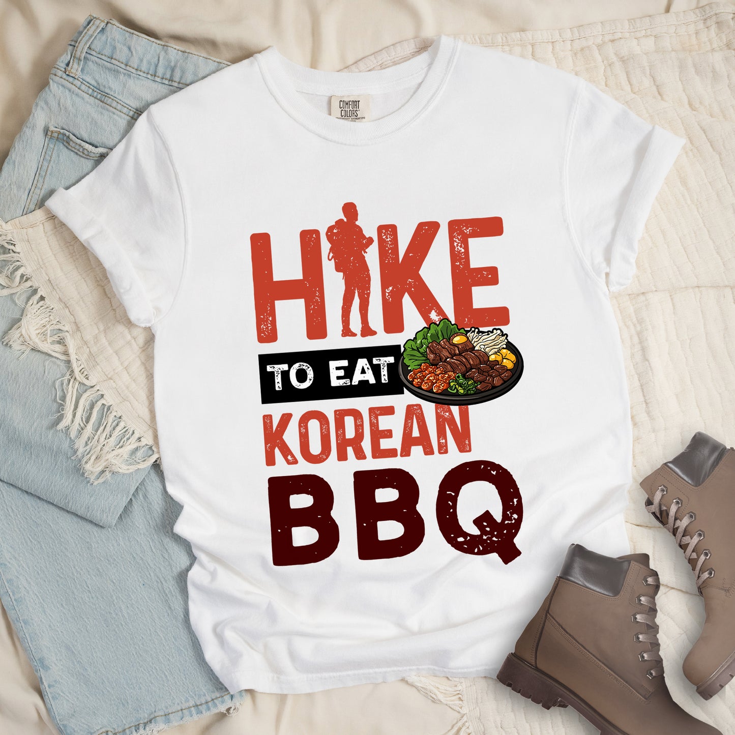 White t-shirt with red and dark red distressed text reading "HIKE TO EAT KOREAN BBQ". "HIKE" appears in large red letters at the top with a red hiker silhouette integrated into the letter "I", "TO EAT" in white text on a black rectangular background in the middle, "KOREAN" in red, and "BBQ" in dark red at the bottom. Features an illustration of Korean barbecue with meat and vegetables on a black plate extending from the black background.