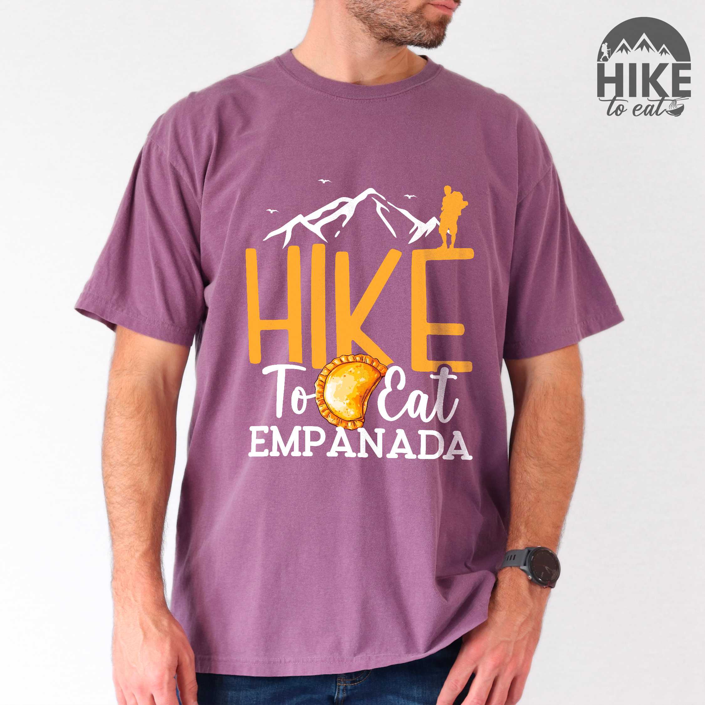 Man wearing berry "Hike to eat empanada" colored vintage tee with bold typography, illustrated freshly baked empanada, mountains, birds, and backpackers for street food enthusiasts.
