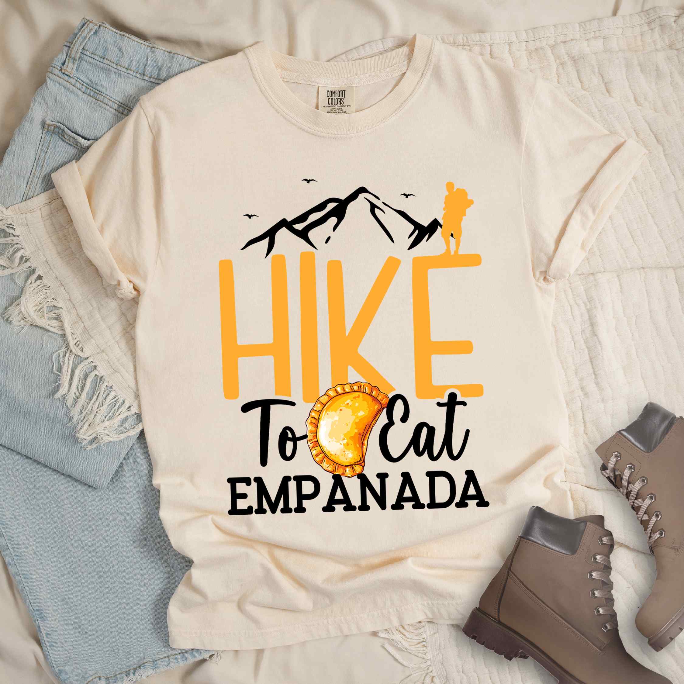 Ivory "Hike to eat empanada" colored vintage tee with bold typography, illustrated freshly baked empanada, mountains, birds, and backpackers for street food enthusiasts.