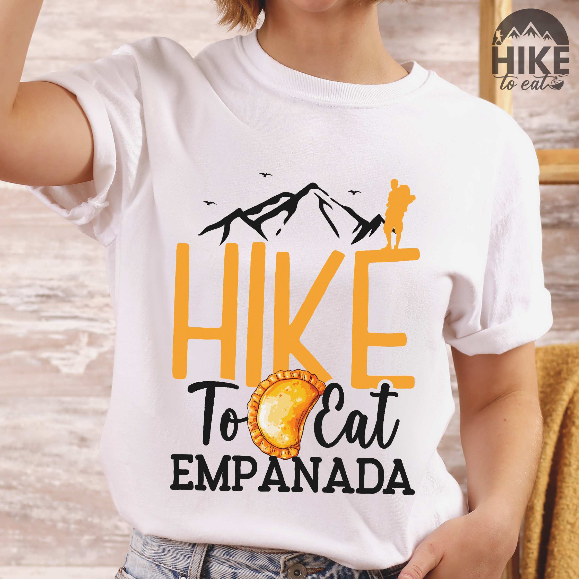 Woman wearing white "Hike to eat empanada" colored vintage tee with bold typography, illustrated freshly baked empanada, mountains, birds, and backpackers for street food enthusiasts.