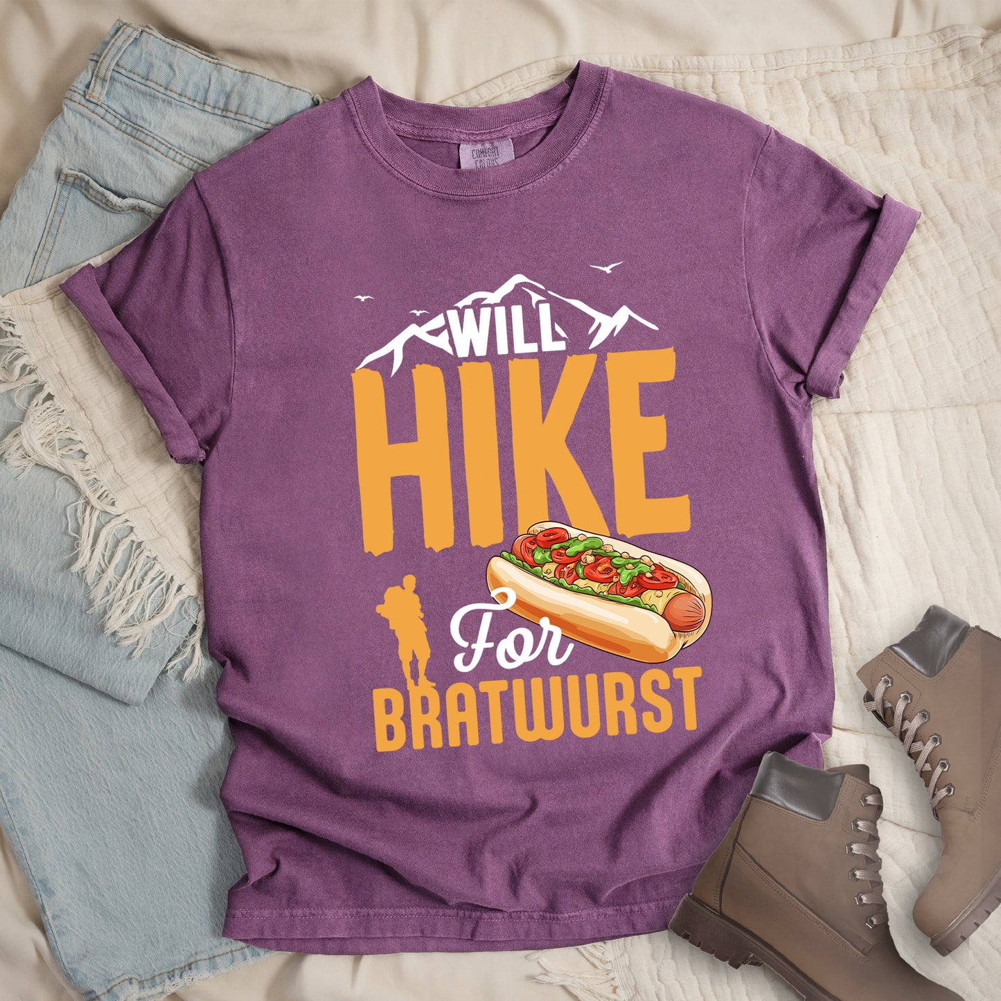 Berry (Dark Violet) t-shirt with white, golden orange, and white text reading "WILL HIKE For BRATWURST". "WILL" appears in white text integrated with mountain outlines at the top, "HIKE" in large golden orange letters in the middle, "For" in white script below it, and "BRATWURST" in golden orange at the bottom. Features an illustration of a bratwurst in a bun with toppings to the right of "For" and a golden orange silhouette of a hiker to the left of "BRATWURST". Small white birds fly near the mountains.