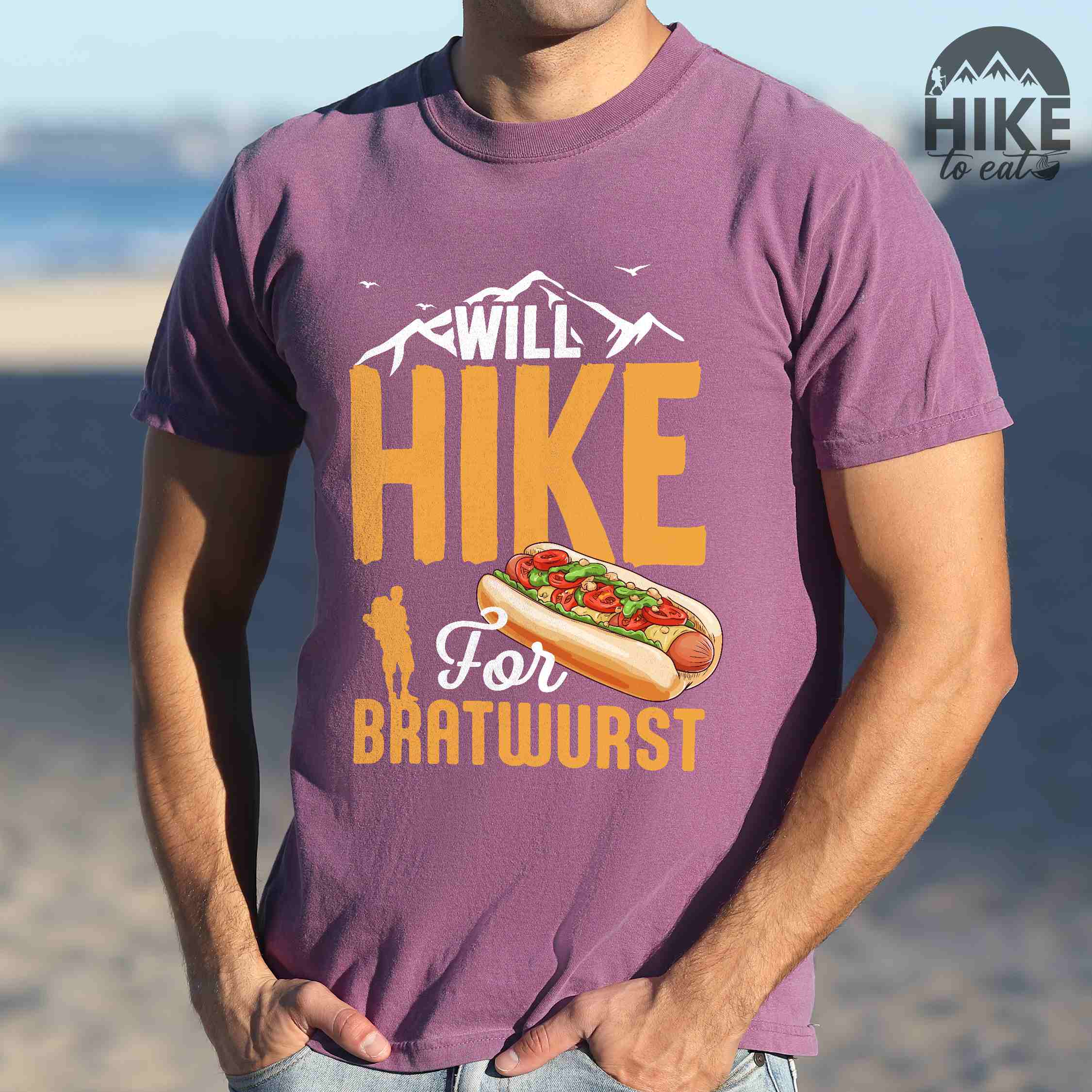 Man wearing berry "Will hike for bratwurst" colored vintage tee with bold typography, illustrated bratwurst in a bun, mountains, birds, and backpackers for street food enthusiasts.