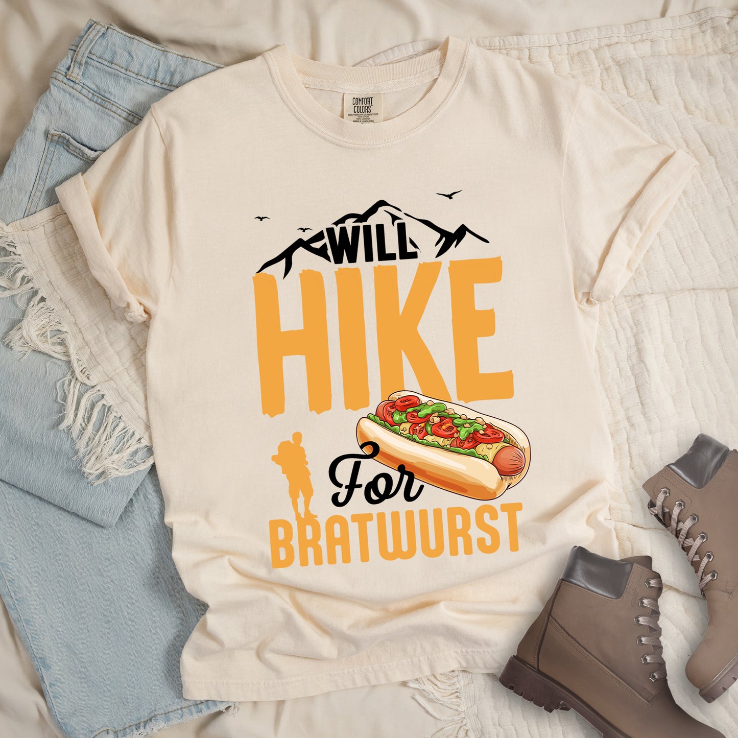 Ivory (Cream White) t-shirt with black, golden orange, and black text reading "WILL HIKE For BRATWURST". "WILL" appears in black text integrated with mountain outlines at the top, "HIKE" in large golden orange letters in the middle, "For" in black script below it, and "BRATWURST" in golden orange at the bottom. Features an illustration of a bratwurst in a bun with toppings to the right of "For" and a golden orange silhouette of a hiker to the left of "BRATWURST". Small black birds fly near the mountains.