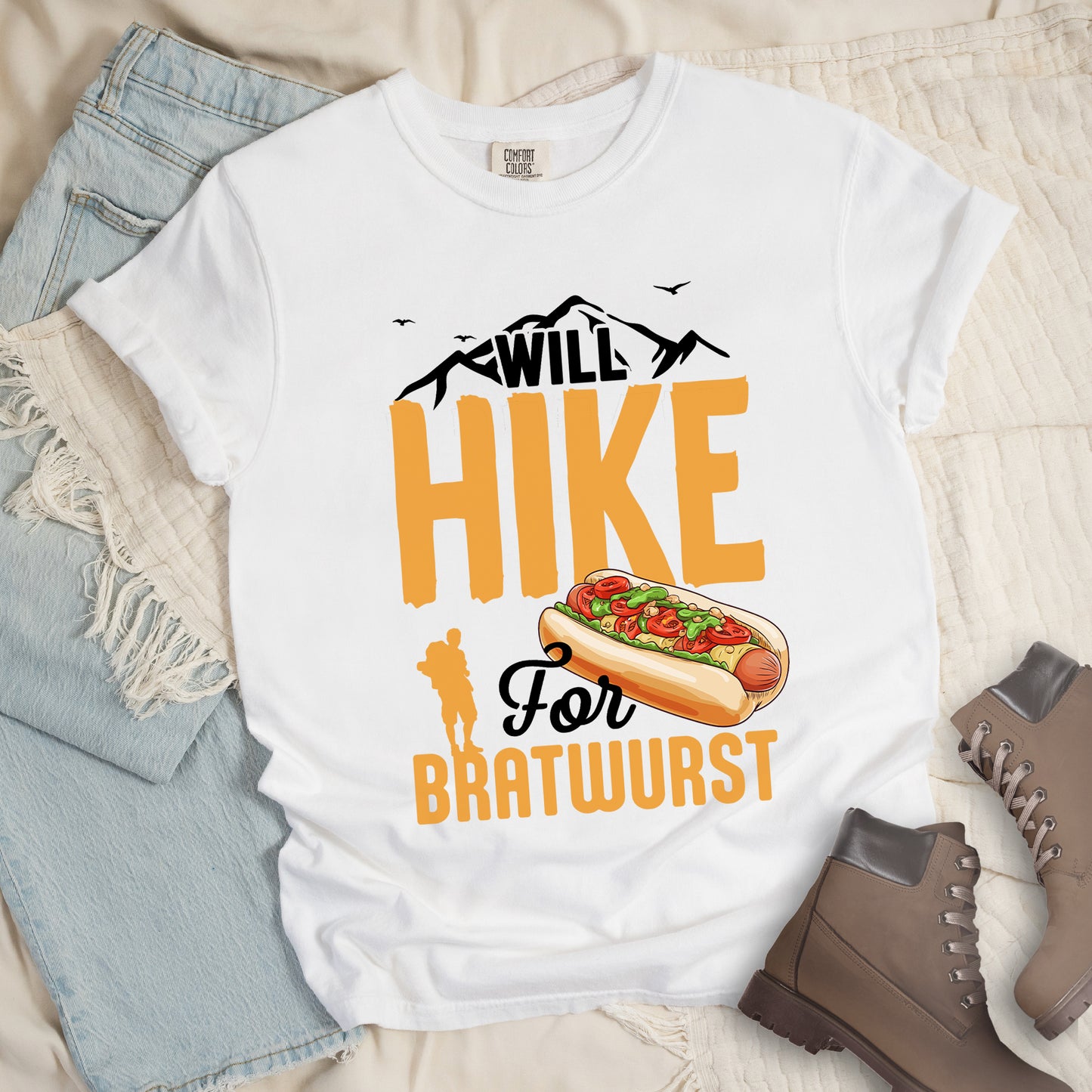 White t-shirt with black, golden orange, and black text reading "WILL HIKE For BRATWURST". "WILL" appears in black text integrated with mountain outlines at the top, "HIKE" in large golden orange letters in the middle, "For" in black script below it, and "BRATWURST" in golden orange at the bottom. Features an illustration of a bratwurst in a bun with toppings to the right of "For" and a golden orange silhouette of a hiker to the left of "BRATWURST". Small black birds fly near the mountains.