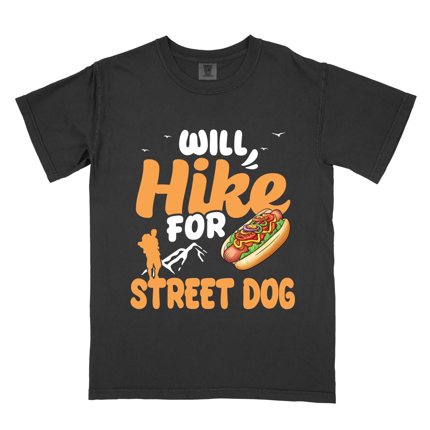 Black t-shirt with orange and white text reading "WILL Hike FOR STREET DOG". "WILL" appears in white at the top with small white birds flying nearby, "Hike" in large orange script in the middle, "FOR" in white below it with small white mountain outlines, and "STREET DOG" in orange at the bottom. Features an illustration of a hot dog with toppings to the right of "FOR" and an orange silhouette of a hiker to the left top of "STREET DOG".