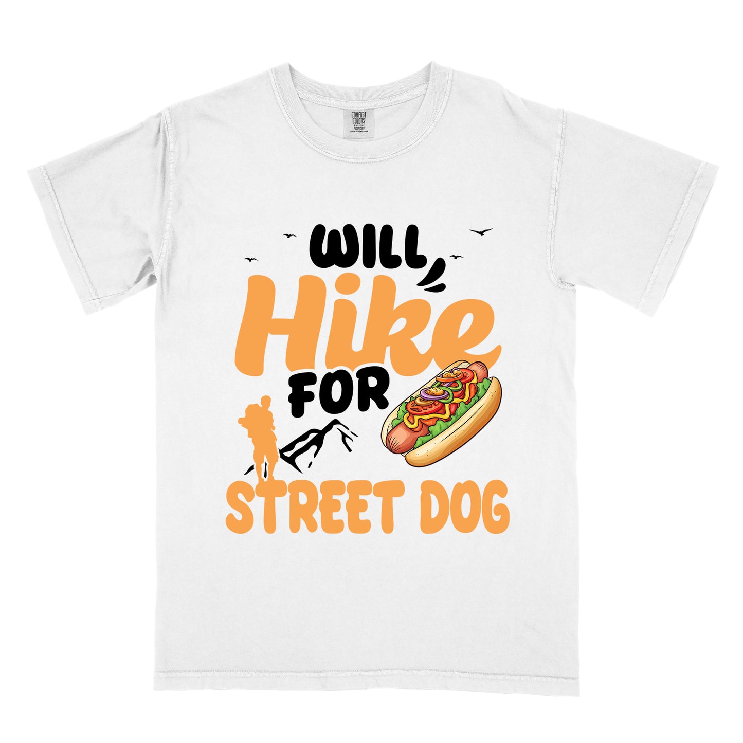 White t-shirt with orange and black text reading "WILL Hike FOR STREET DOG". "WILL" appears in black at the top with small black birds flying nearby, "Hike" in large orange script in the middle, "FOR" in black below it with small black mountain outlines, and "STREET DOG" in orange at the bottom. Features an illustration of a hot dog with toppings to the right of "FOR" and an orange silhouette of a hiker to the left top of "STREET DOG".