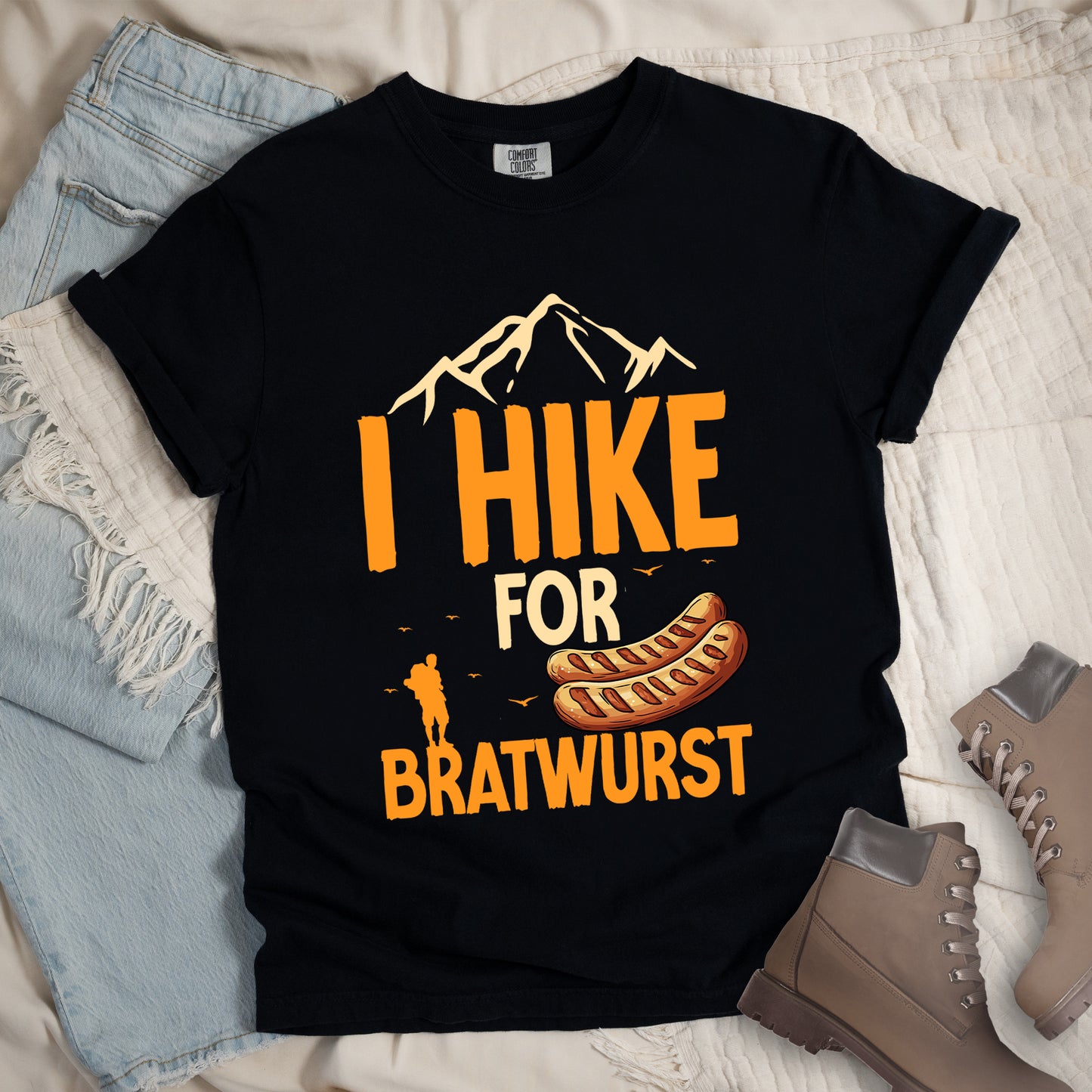 Black t-shirt with orange and beige text reading "I HIKE FOR BRATWURST". "I HIKE" appears in large orange letters at the top with beige mountain outlines above it, "FOR" in beige text in the middle, and "BRATWURST" in orange at the bottom. Features an illustration of two bratwurst sausages and a small orange hiker silhouette with birds flying nearby above the word "BRATWURST".