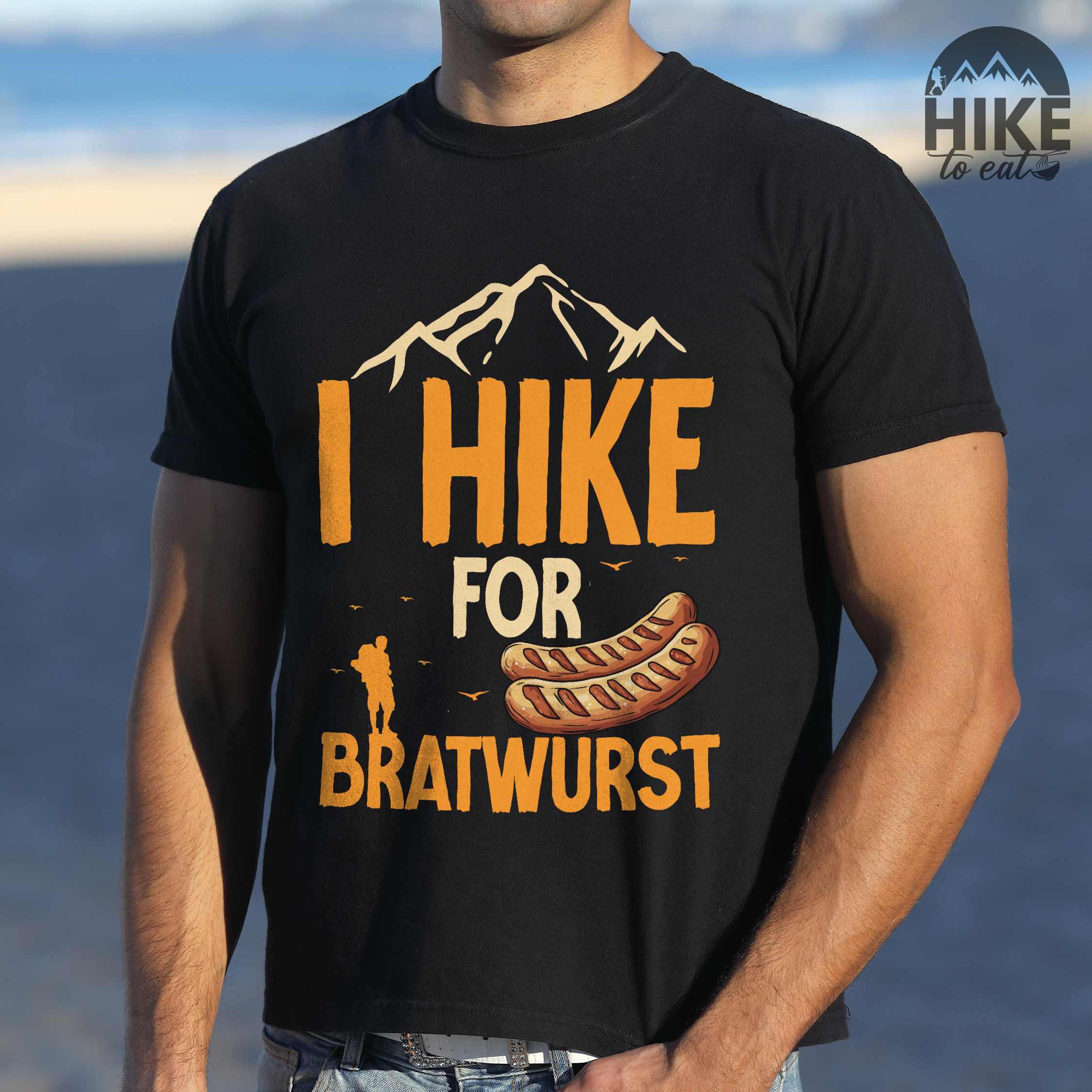 Man wearing black "I hike for bratwurst" colored vintage tee with bold typography, illustrated bratwurst, mountains, birds, and backpackers for street dog lovers.