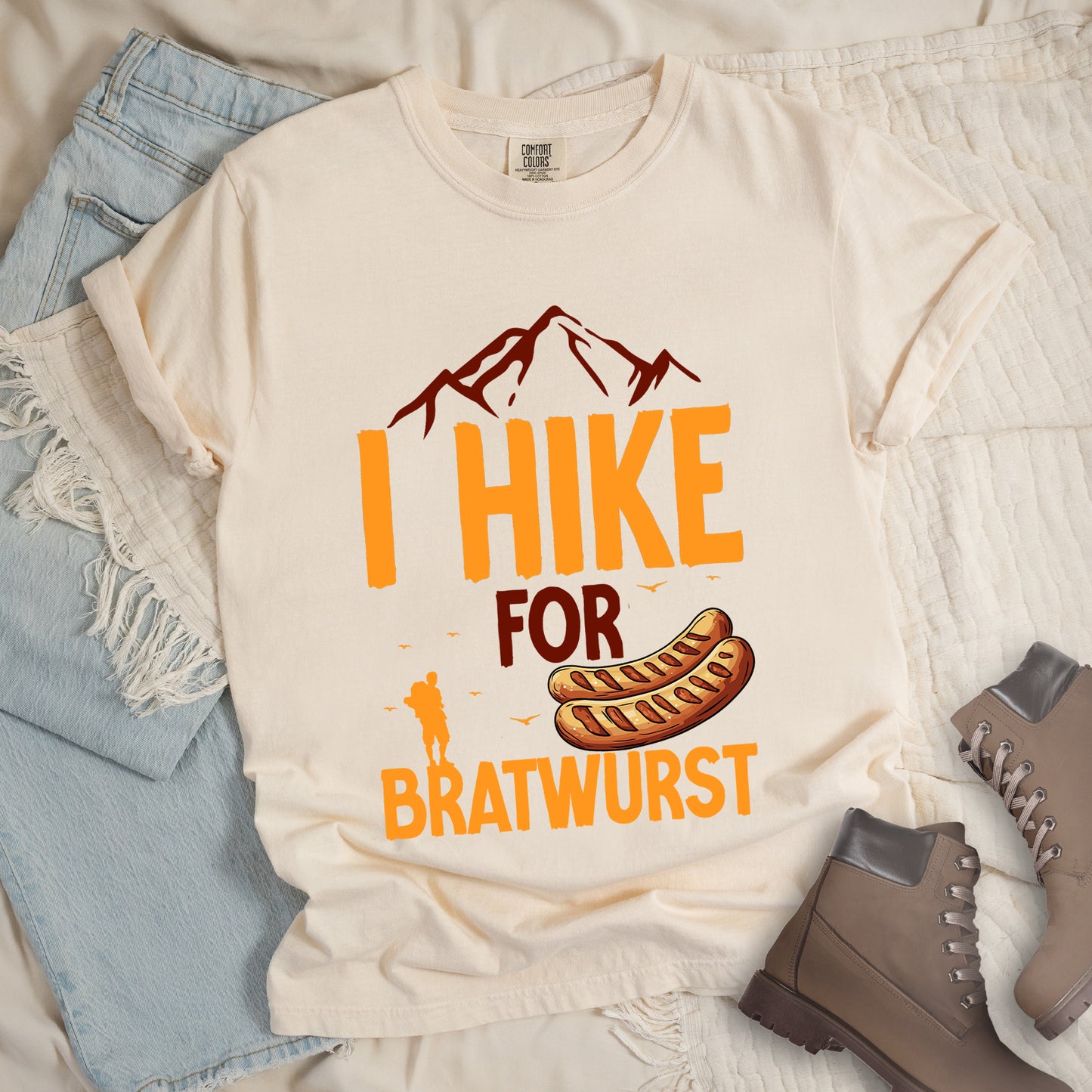 Ivory (Cream White) t-shirt with orange and brown text reading "I HIKE FOR BRATWURST". "I HIKE" appears in large orange letters at the top with brown mountain outlines above it, "FOR" in brown text in the middle, and "BRATWURST" in orange at the bottom. Features an illustration of two bratwurst sausages and a small orange hiker silhouette with birds flying nearby above the word "BRATWURST".