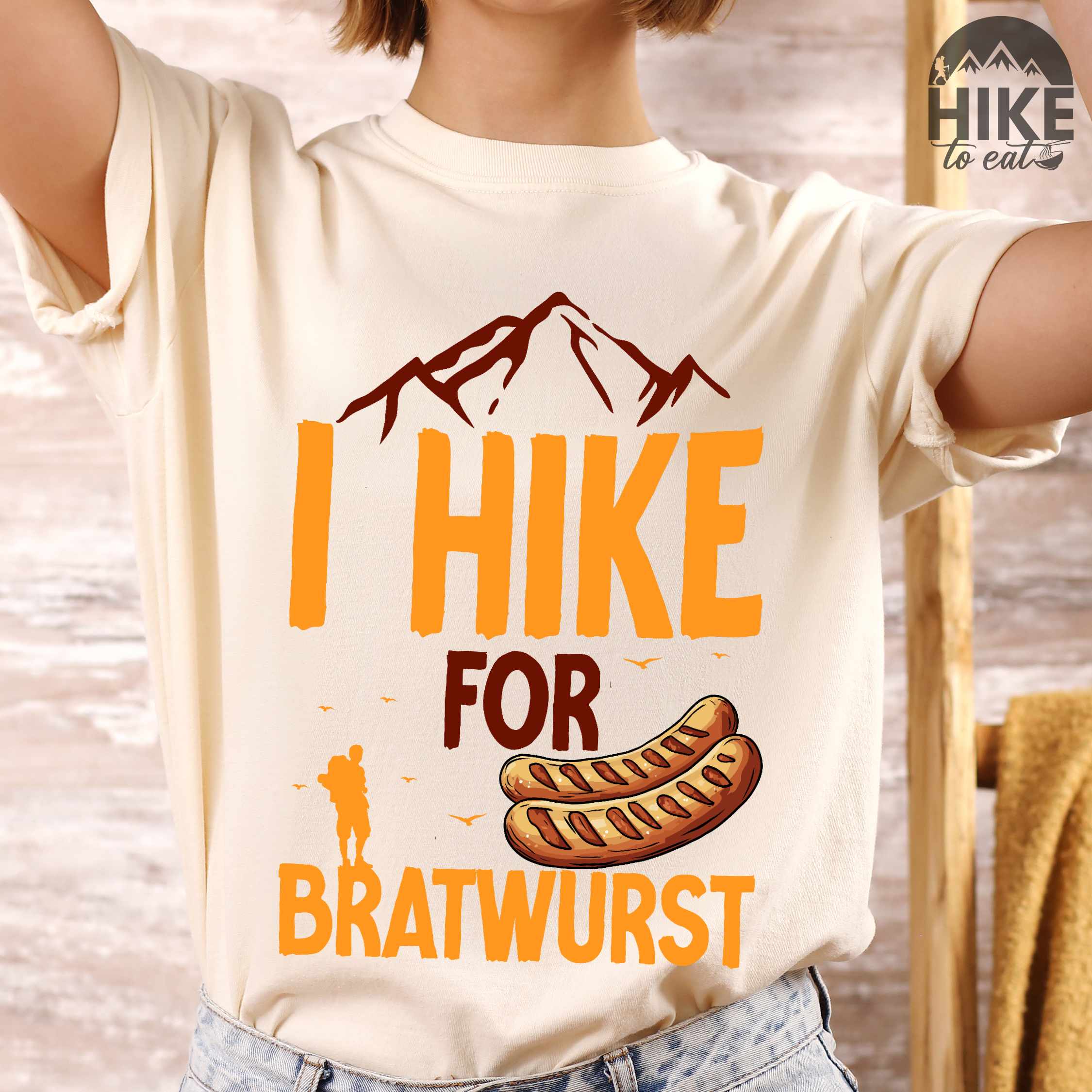 Woman wearing ivory "I hike for bratwurst" colored vintage tee with bold typography, illustrated bratwurst, mountains, birds, and backpackers for street dog lovers.
