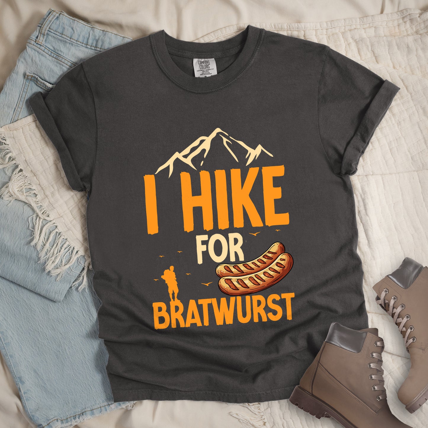 Pepper (Dark Gray) t-shirt with orange and beige text reading "I HIKE FOR BRATWURST". "I HIKE" appears in large orange letters at the top with beige mountain outlines above it, "FOR" in beige text in the middle, and "BRATWURST" in orange at the bottom. Features an illustration of two bratwurst sausages and a small orange hiker silhouette with birds flying nearby above the word "BRATWURST".