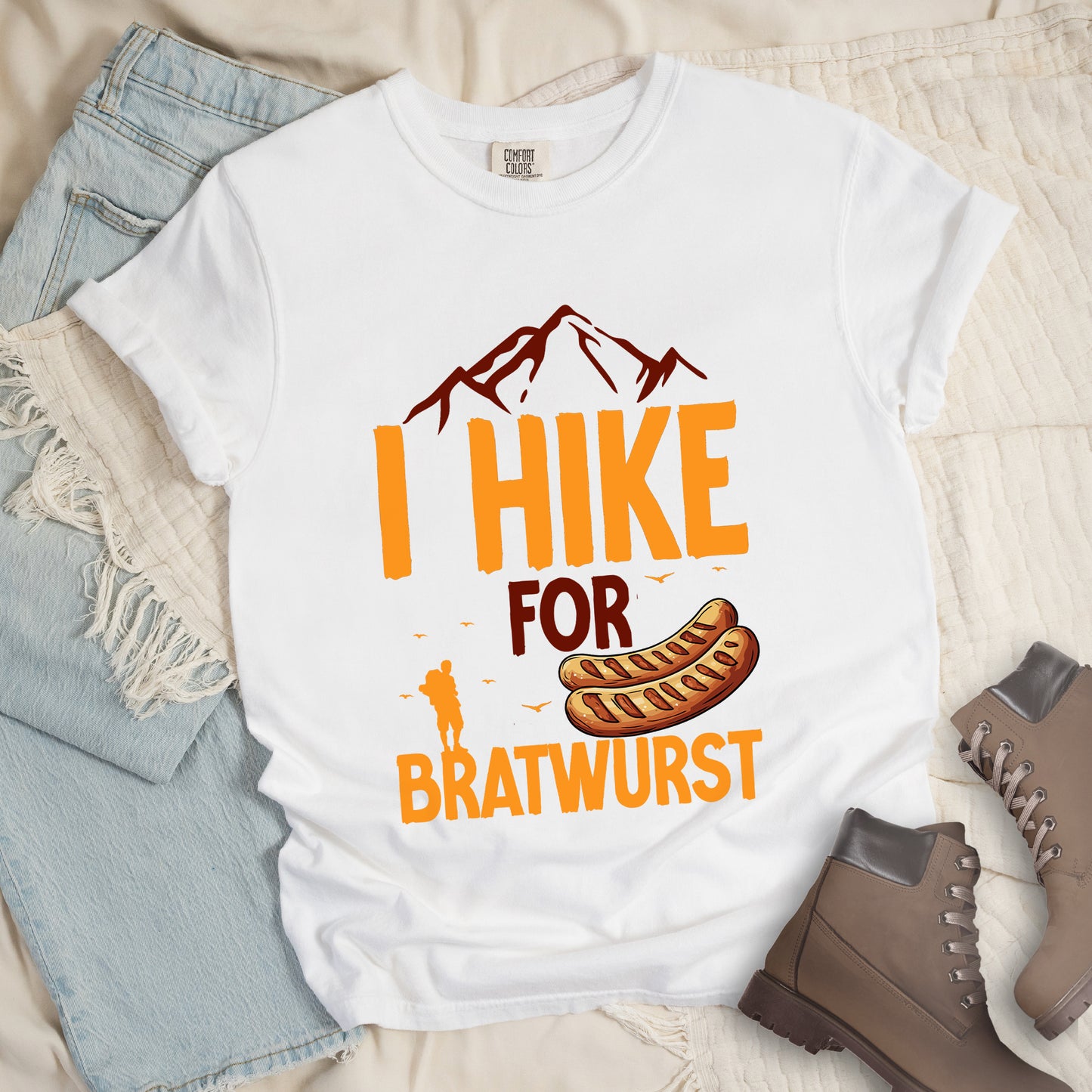 White t-shirt with orange and brown text reading "I HIKE FOR BRATWURST". "I HIKE" appears in large orange letters at the top with brown mountain outlines above it, "FOR" in brown text in the middle, and "BRATWURST" in orange at the bottom. Features an illustration of two bratwurst sausages and a small orange hiker silhouette with birds flying nearby above the word "BRATWURST".