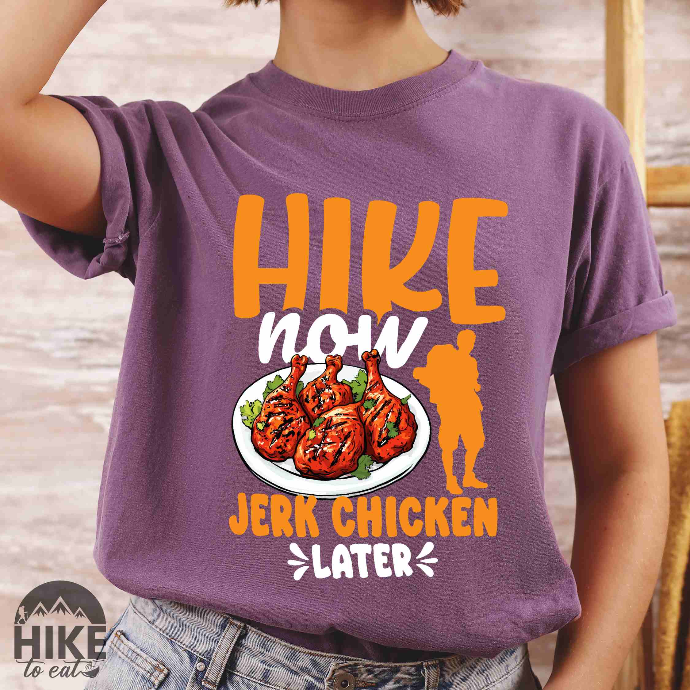 Woman wearing berry "hike now jerk chicken later" colored vintage tee with bold typography, illustrated jerk chicken on a plate, mountains, birds, and backpackers for bbq chicken lovers.