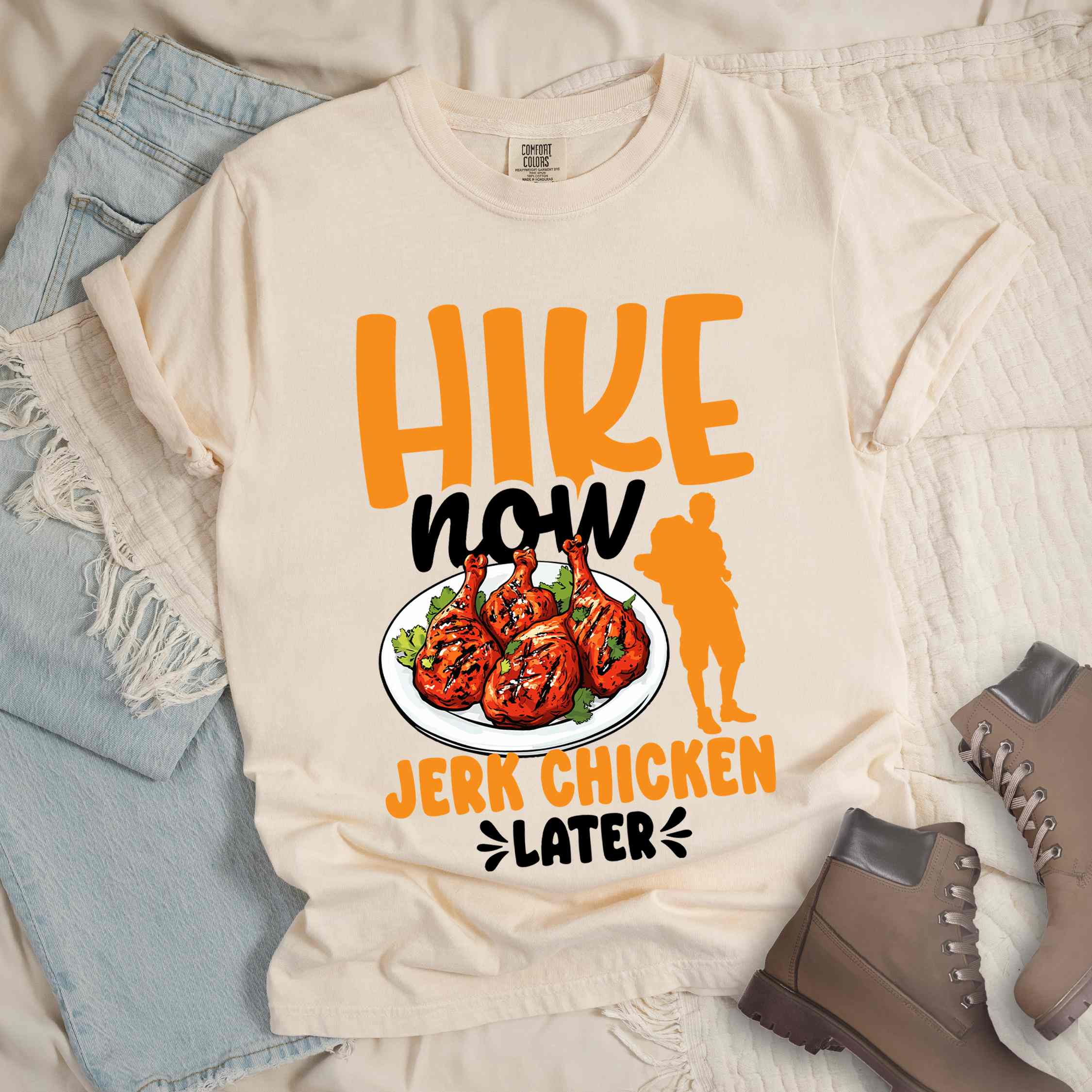 Ivory "hike now jerk chicken later" colored vintage tee with bold typography, illustrated jerk chicken on a plate, mountains, birds, and backpackers for bbq chicken lovers.