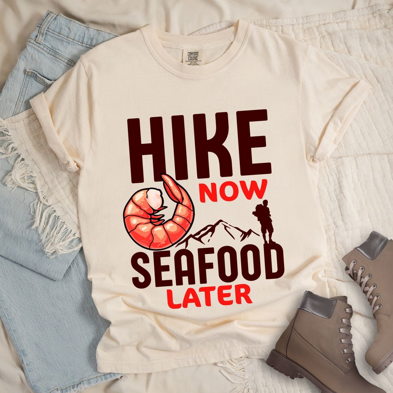 Funny hiking motivation shirt for seafood lovers featuring bold NOW LATER typography, realistic shrimp graphic, mountain landscape illustration, and hiker silhouette on premium outdoor adventure tee perfect gift for trail enthusiasts who love fresh ocean food in ivory (beige) layed flat on boots and jeans