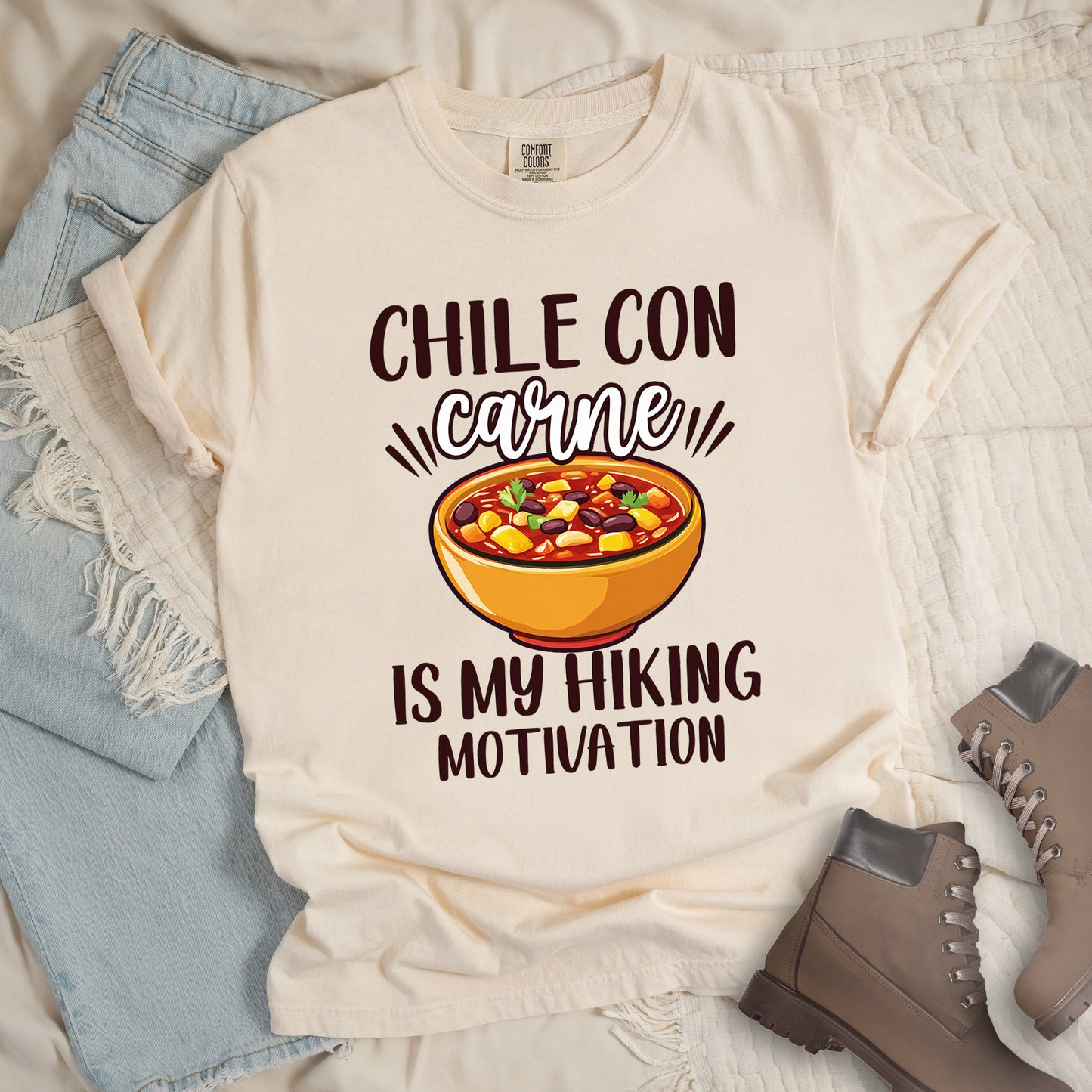 Ivory (Cream White) t-shirt with black text reading "CHILE CON carne IS MY HIKING MOTIVATION." The word "carne" appears in stylized script. In the center is a colorful illustration of a bowl of chili con carne showing beans, meat, corn, and vegetables in an orange bowl. The shirt features a classic crew neck and casual rolled sleeves, styled alongside light wash denim and brown hiking boots.